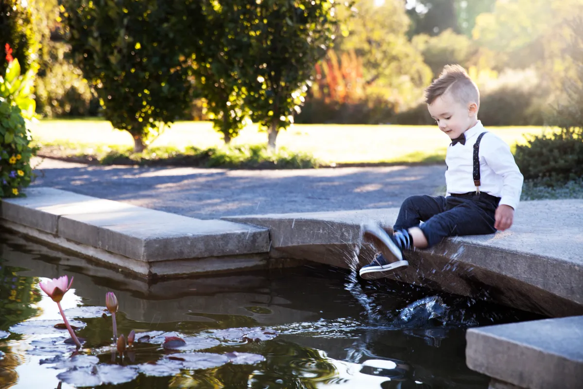 Young boy dressed in formal attire, sitting on the edge of a stone fountain, splashing water with his feet, in a garden or park on a sunny day.
