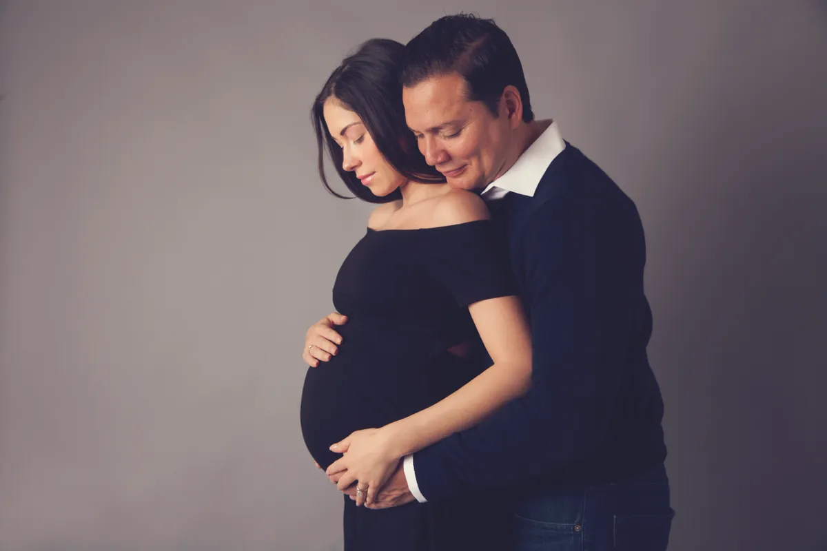 A pregnant woman in a black dress and a man in a dark suit with a white shirt embracing against a plain gray background.