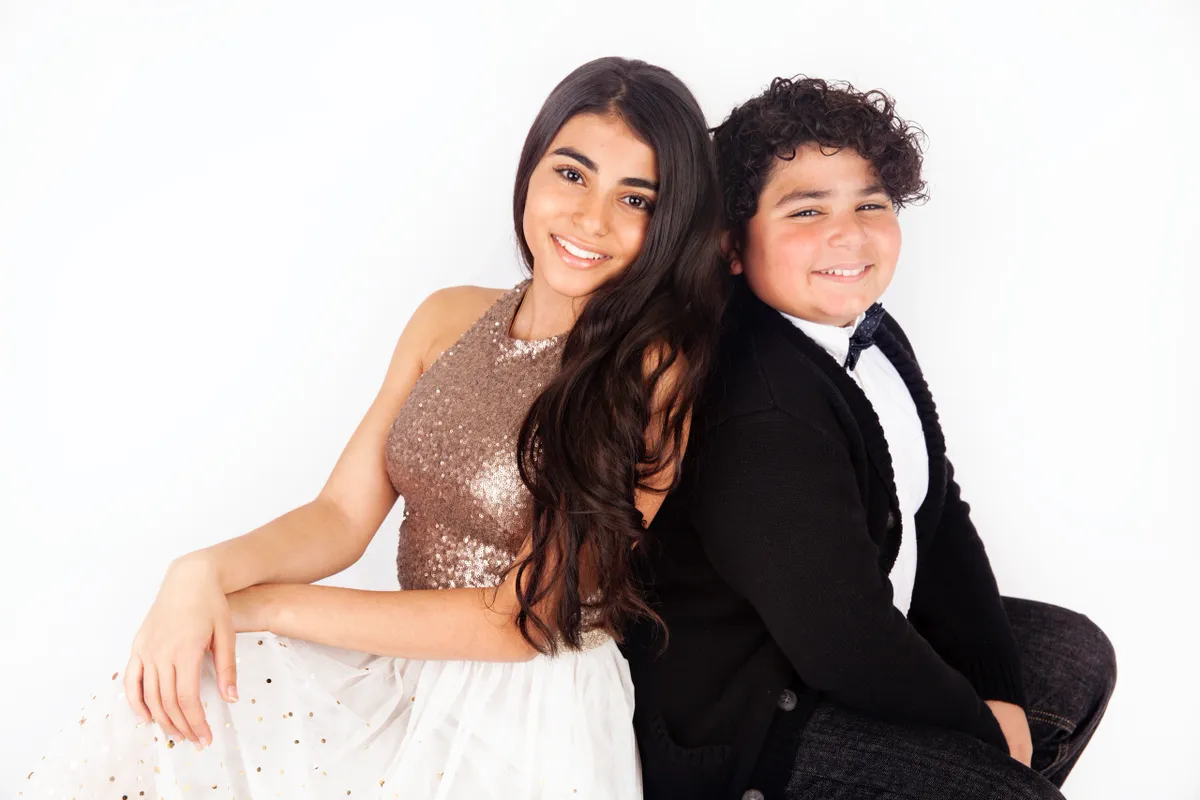 A young woman and young boy sitting back to back and smiling at the camera against a plain white background.