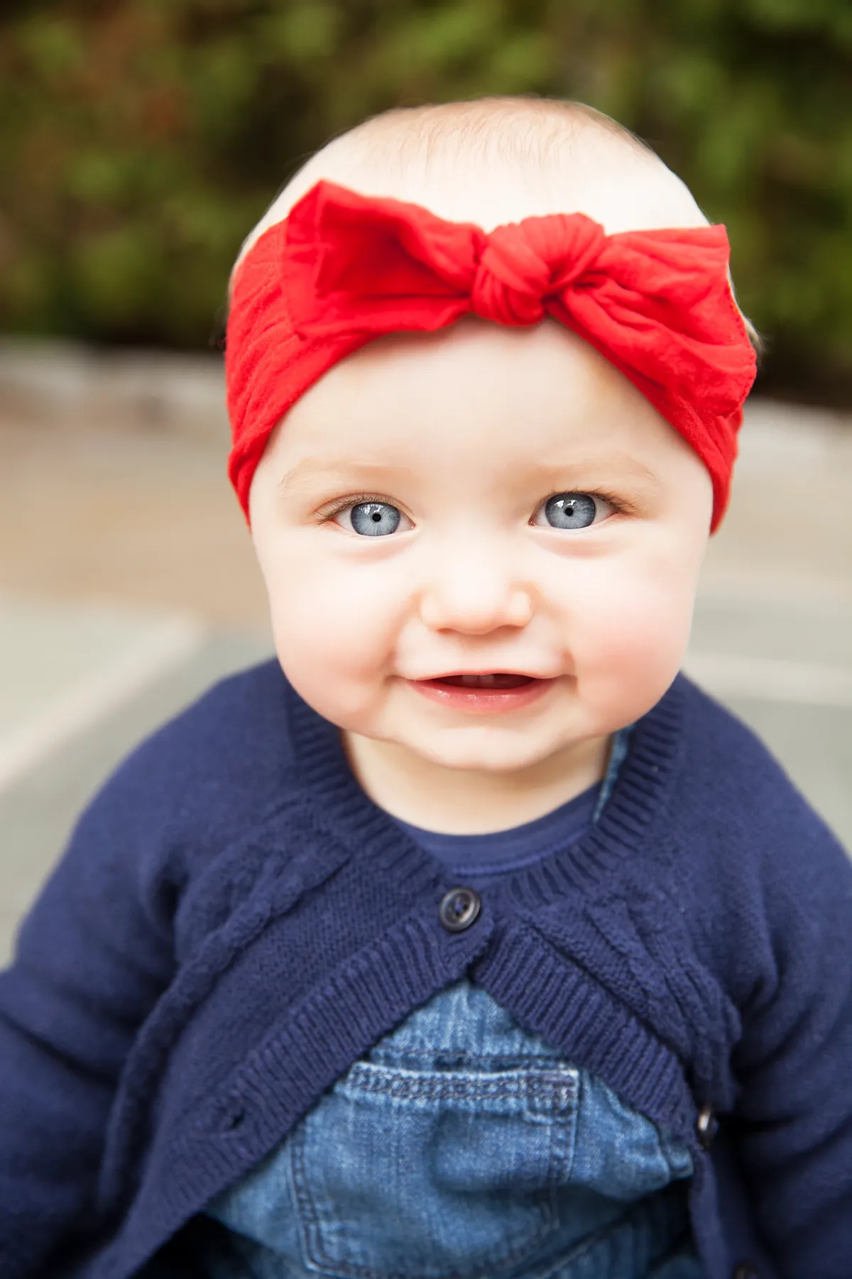 Close-up of a smiling baby girl with blue eyes wearing a red headband, a navy blue cardigan, and denim overalls, outdoors with blurred trees in the background.