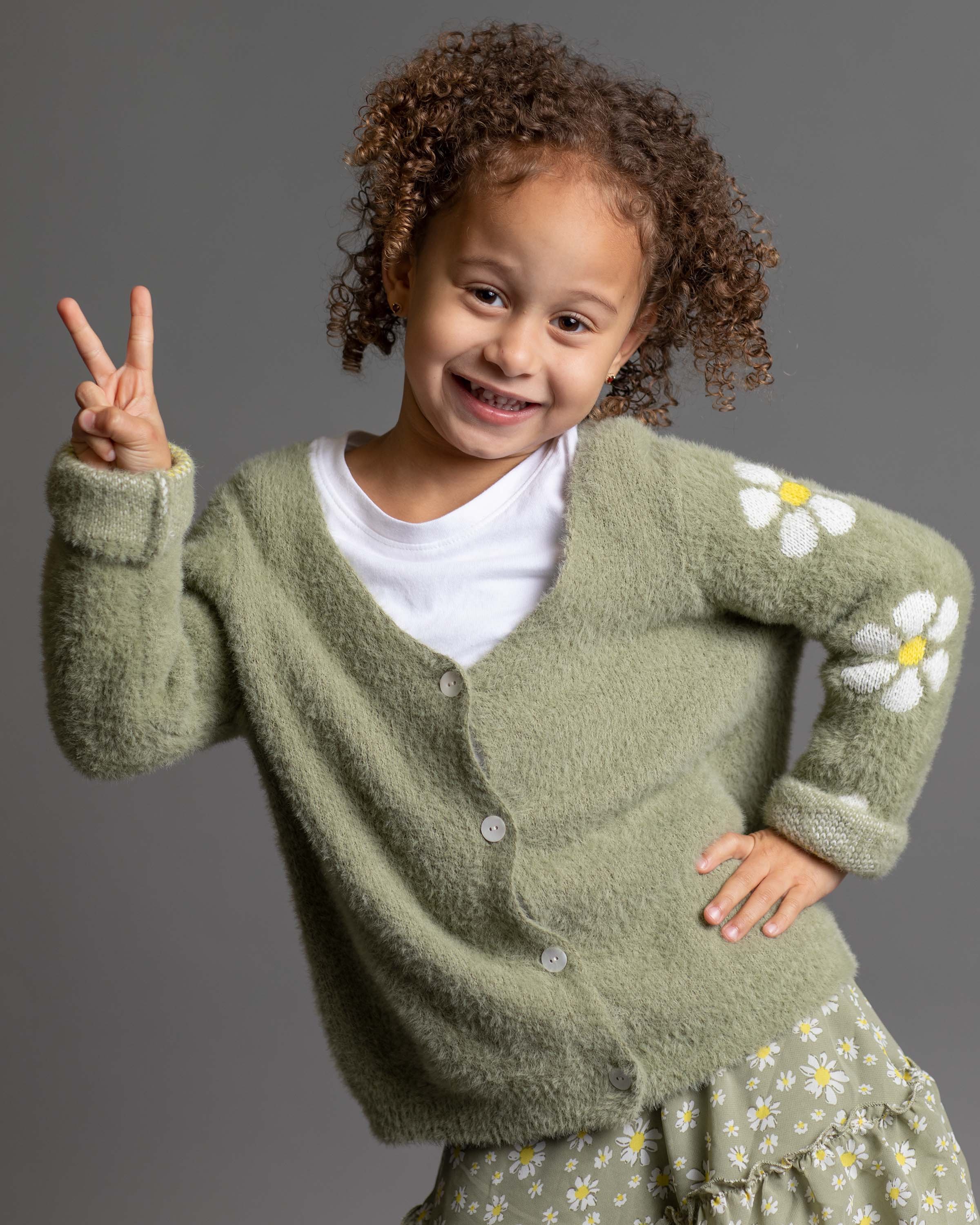 A young girl with curly hair smiling and making a peace sign with her right hand, wearing a green cardigan with daisies on the sleeve and a matching skirt, standing against a plain gray background.