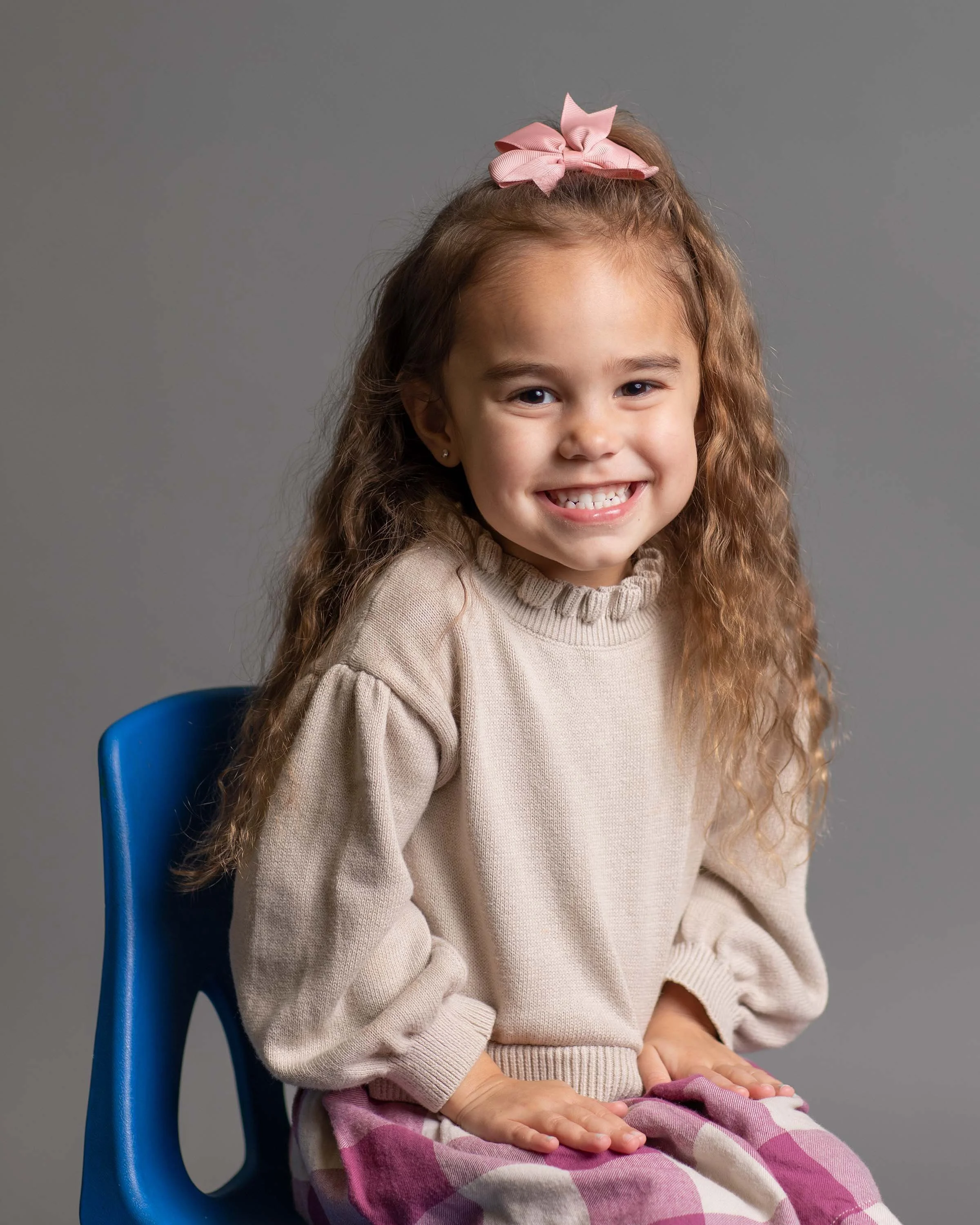A young girl with curly hair wearing a pink bow, beige sweater, and plaid skirt sitting on a blue chair against a gray background, smiling at the camera.