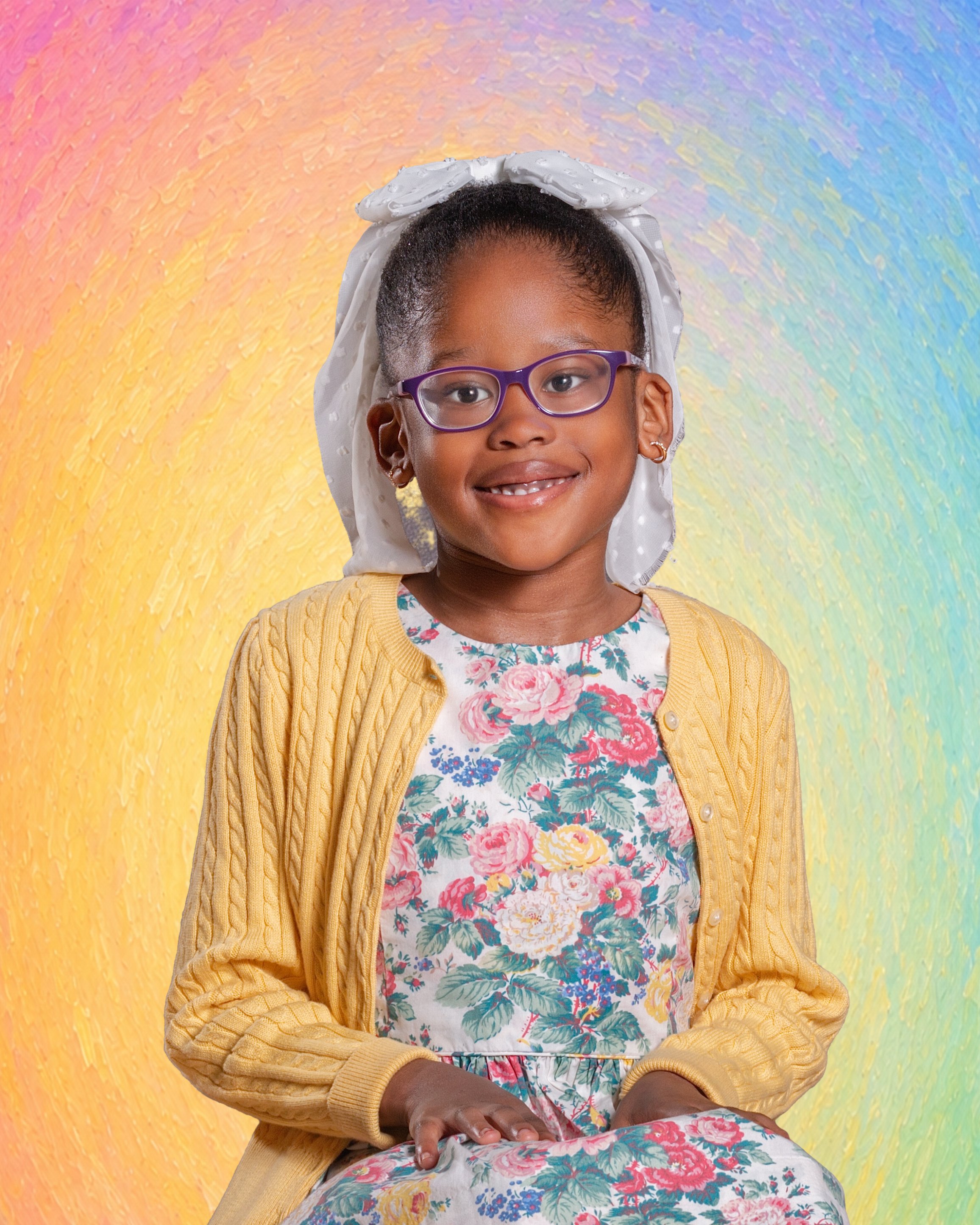 A smiling young girl with curly hair, wearing a beige sweater and checkered pink pajama pants, sitting on a blue chair against a gray background, with a pink bow in her hair.