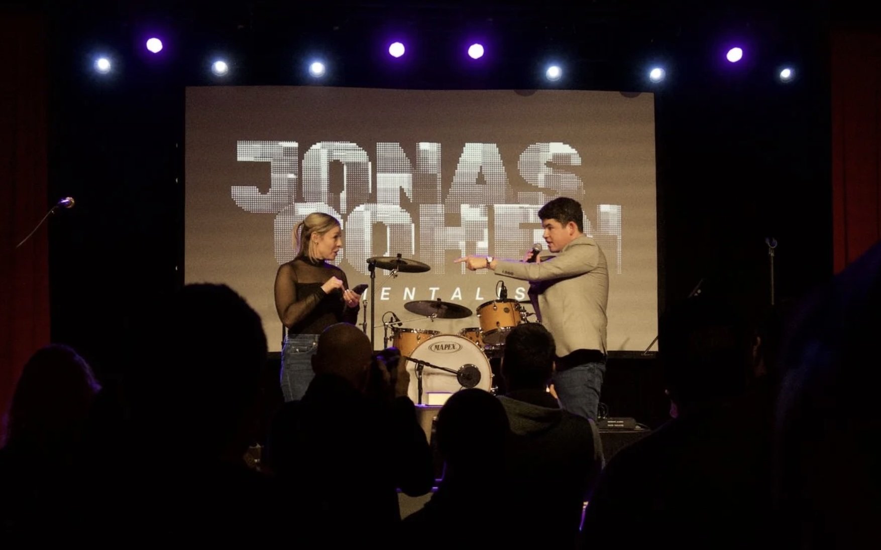 Two people on stage, a woman and a man, talking and pointing at each other, behind a drum set, with a large screen in the background displaying the text 'JONAS BROTHERS' and 'ENTALS'.