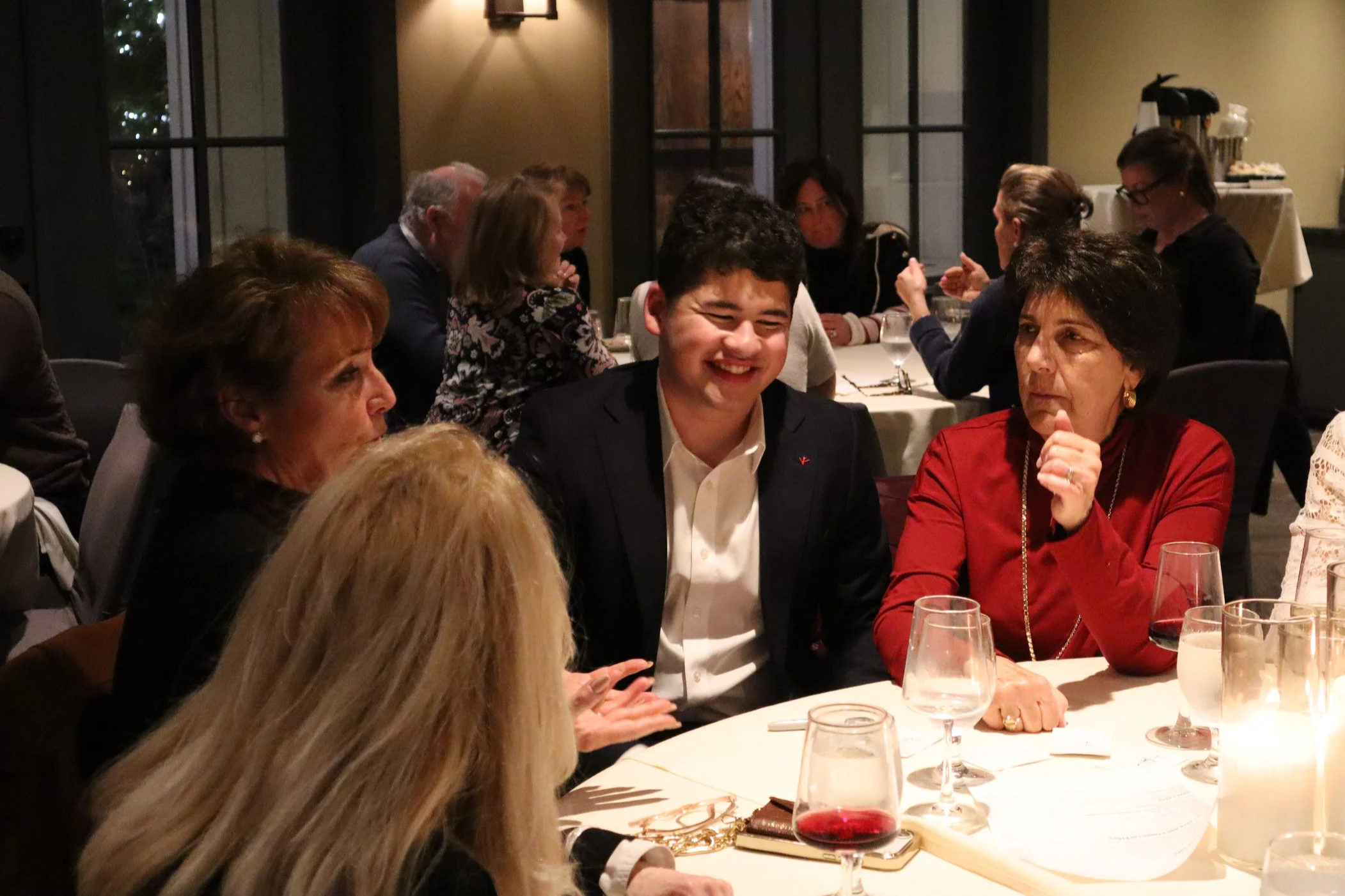 A group of people sitting around a dinner table in a dimly lit restaurant, engaged in conversation. One young man in a black blazer and white shirt is smiling, while a woman in a red top appears to be listening attentively. Other guests are visible in the background.