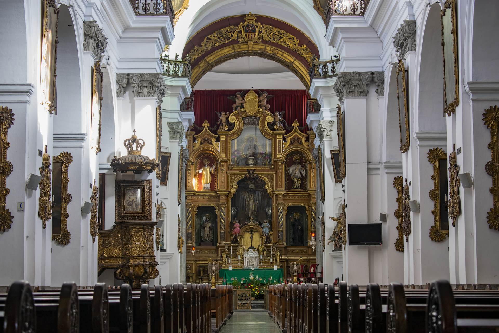 Interior de una iglesia con un altar dorado y detalles religiosos en el fondo, paredes blancas adornadas con cuadros y esculturas.