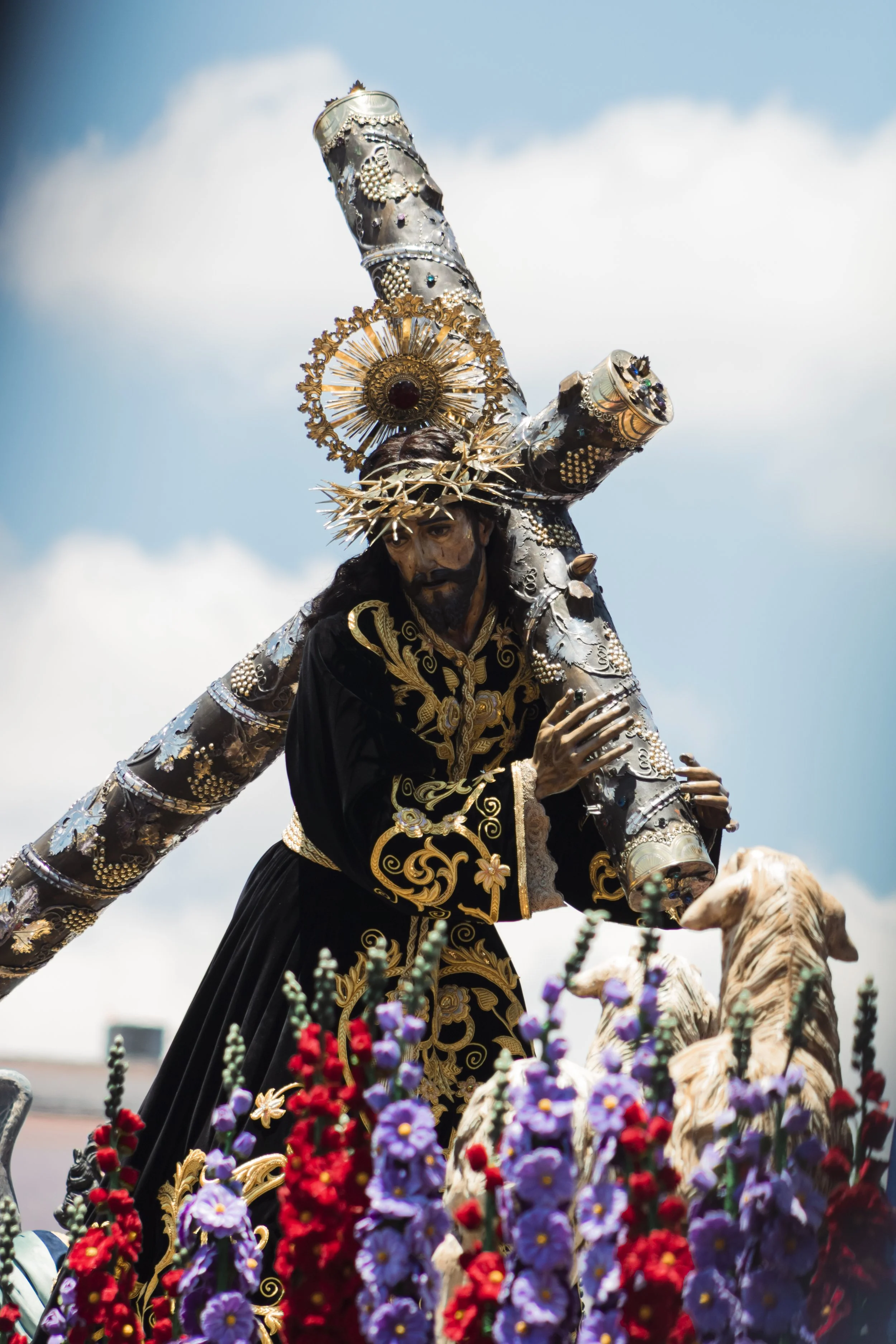 Escultura religiosa de Jesús cargando una cruz, vestida con ropas negras y doradas, con una corona de espinas, rodeada de flores de colores.
