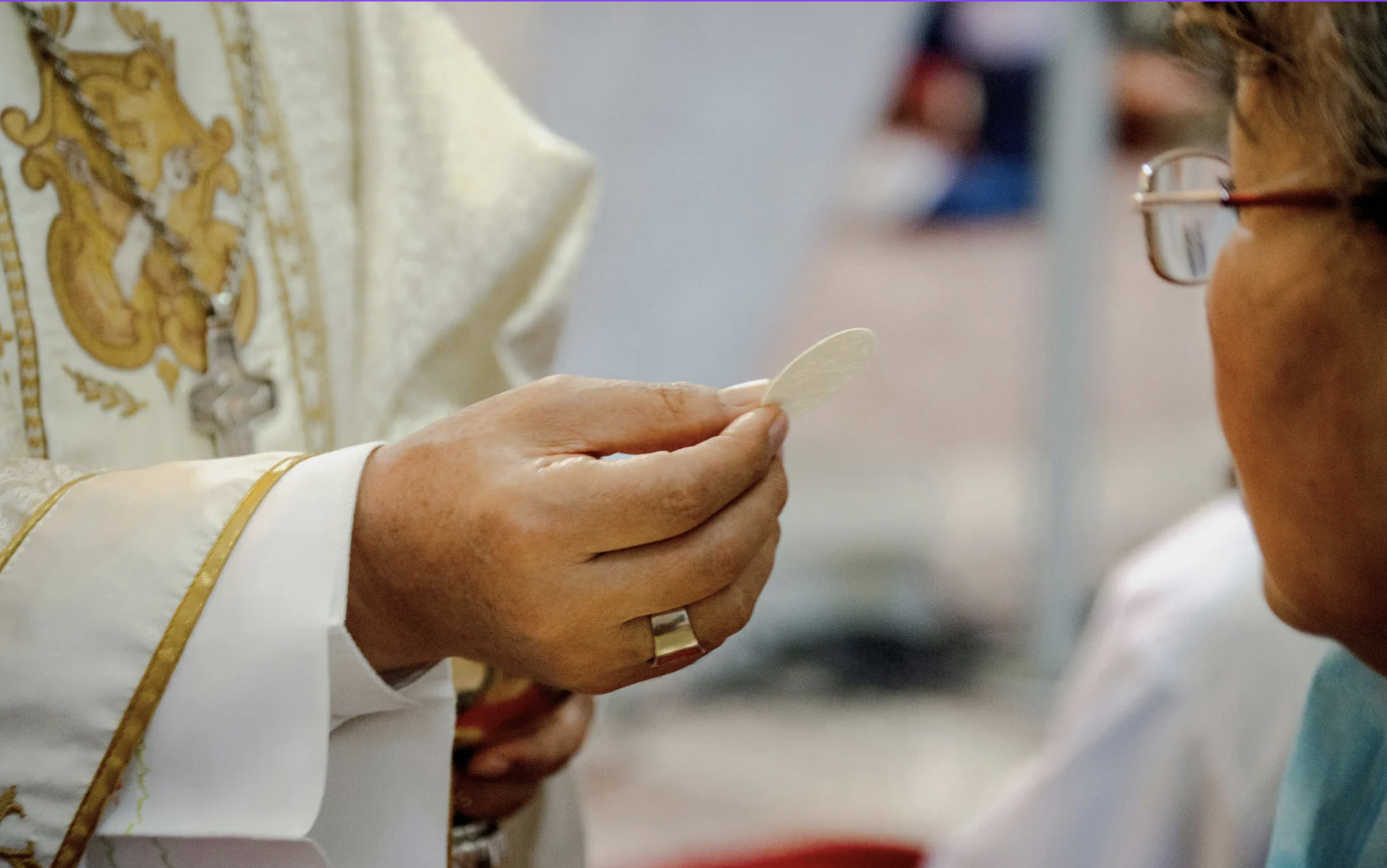 Un sacerdote con vestimenta eclesiástica sosteniendo una hostia en una mano, en una ceremonia religiosa con una mujer que lleva lentes.