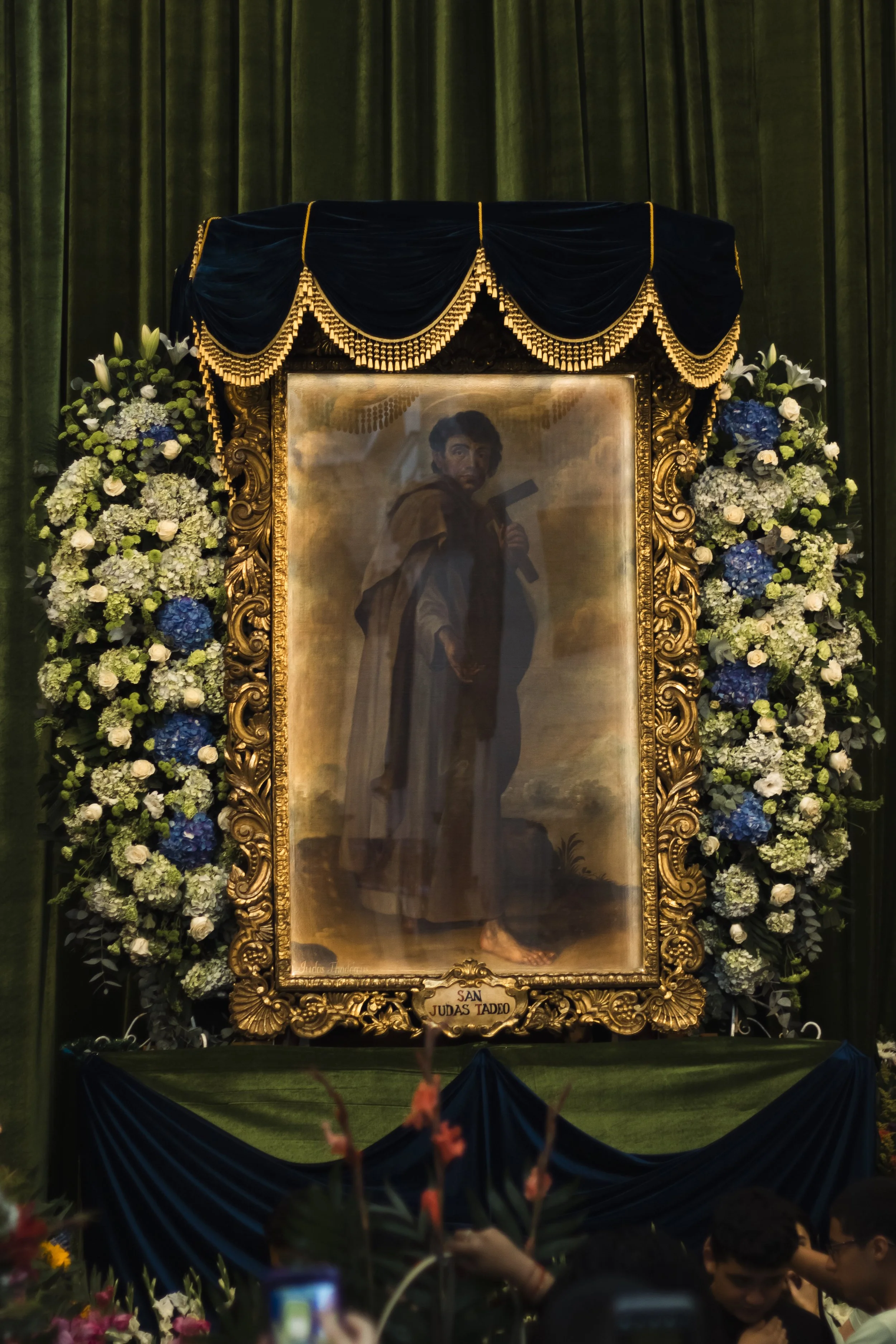 Retrato de San Judas Tadeo rodeado de arreglos florales en un altar con cortinas verdes y decoraciones doradas.