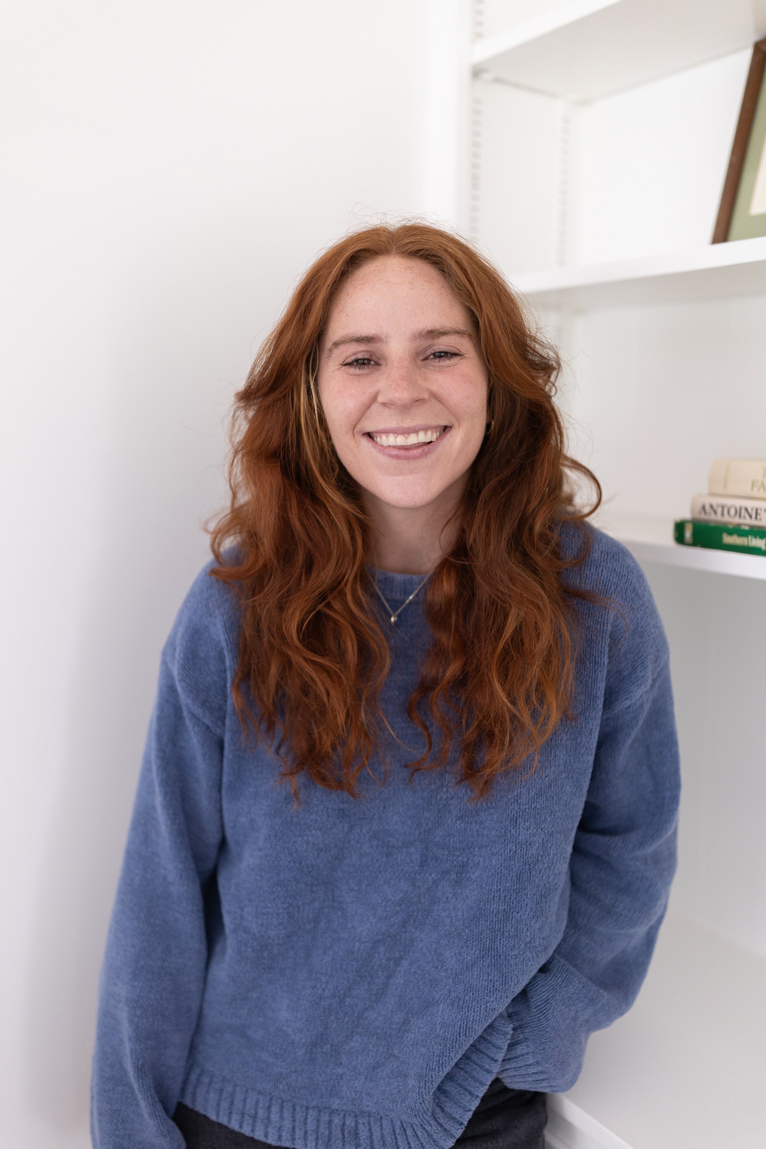 Hayley Hinde-Pettinger smiling at the camera, wearing a blue sweater, standing in front of a white wall and shelves.
