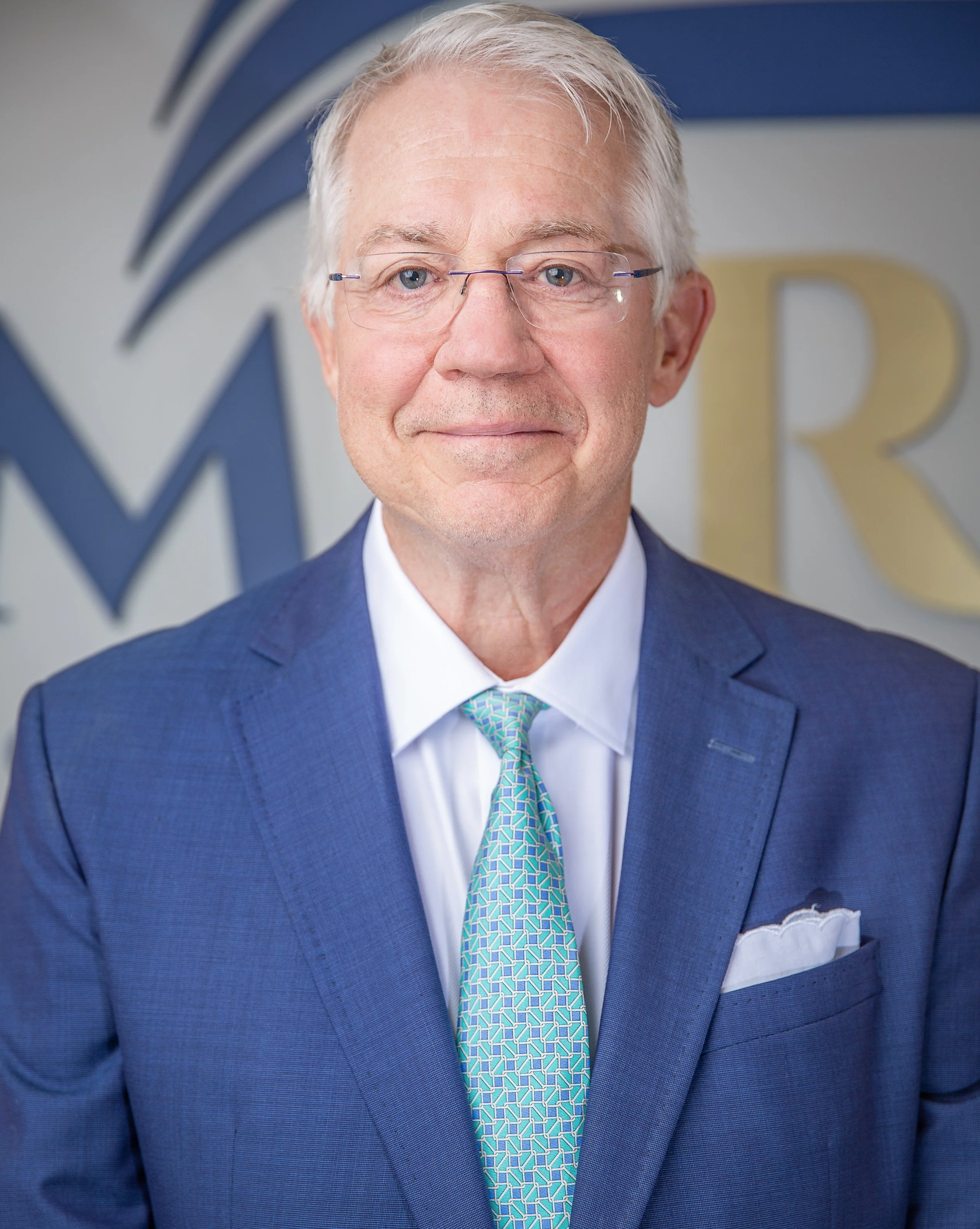 Executive portrait of an older businessman with short white hair and glasses wearing a blue suit, light blue shirt, and patterned teal tie. He stands confidently in front of a modern wall with large architectural lettering in the background.