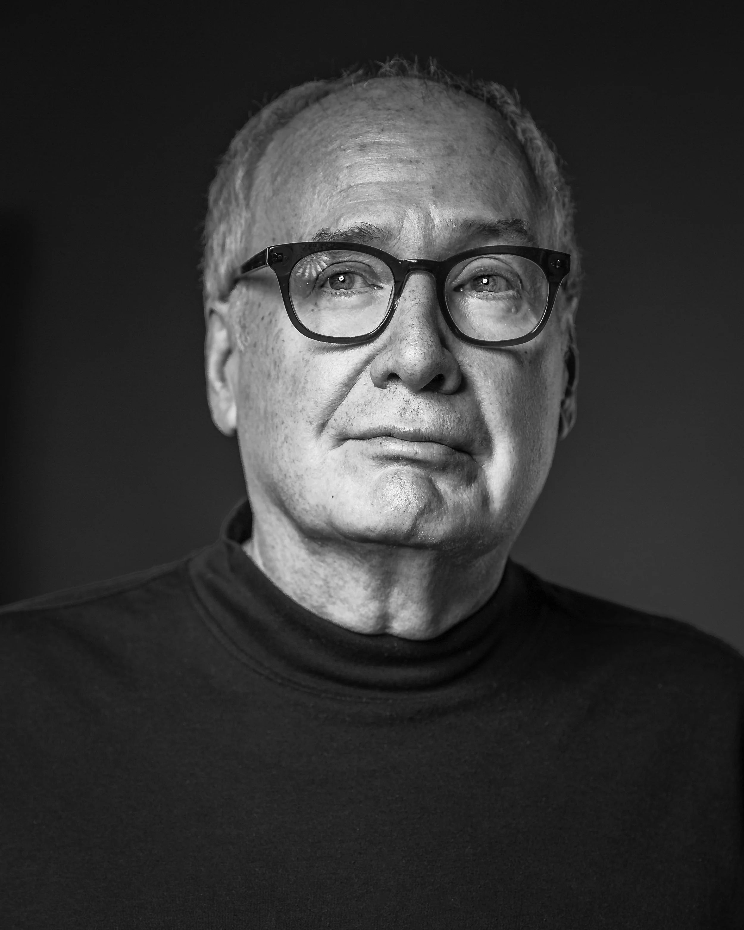 Black and white portrait of an older man with short hair and glasses, wearing a dark turtleneck, looking slightly upward with a thoughtful expression against a plain background.