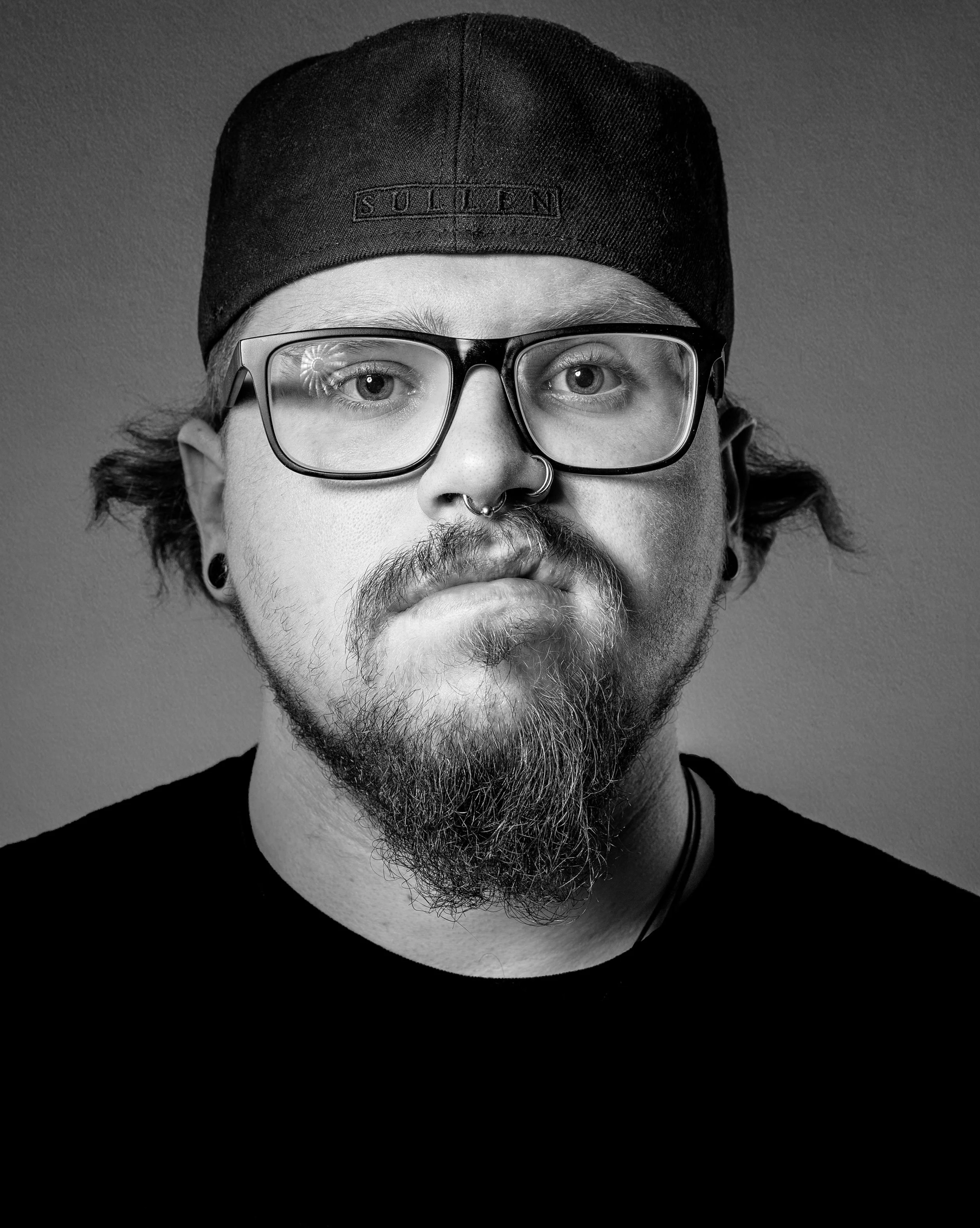 Artist portrait of tattoo artist Benny Zickmanis in black and white. Wearing glasses, a backward cap, and a dark shirt, he looks calmly into the camera, reflecting his distinctive creative style.