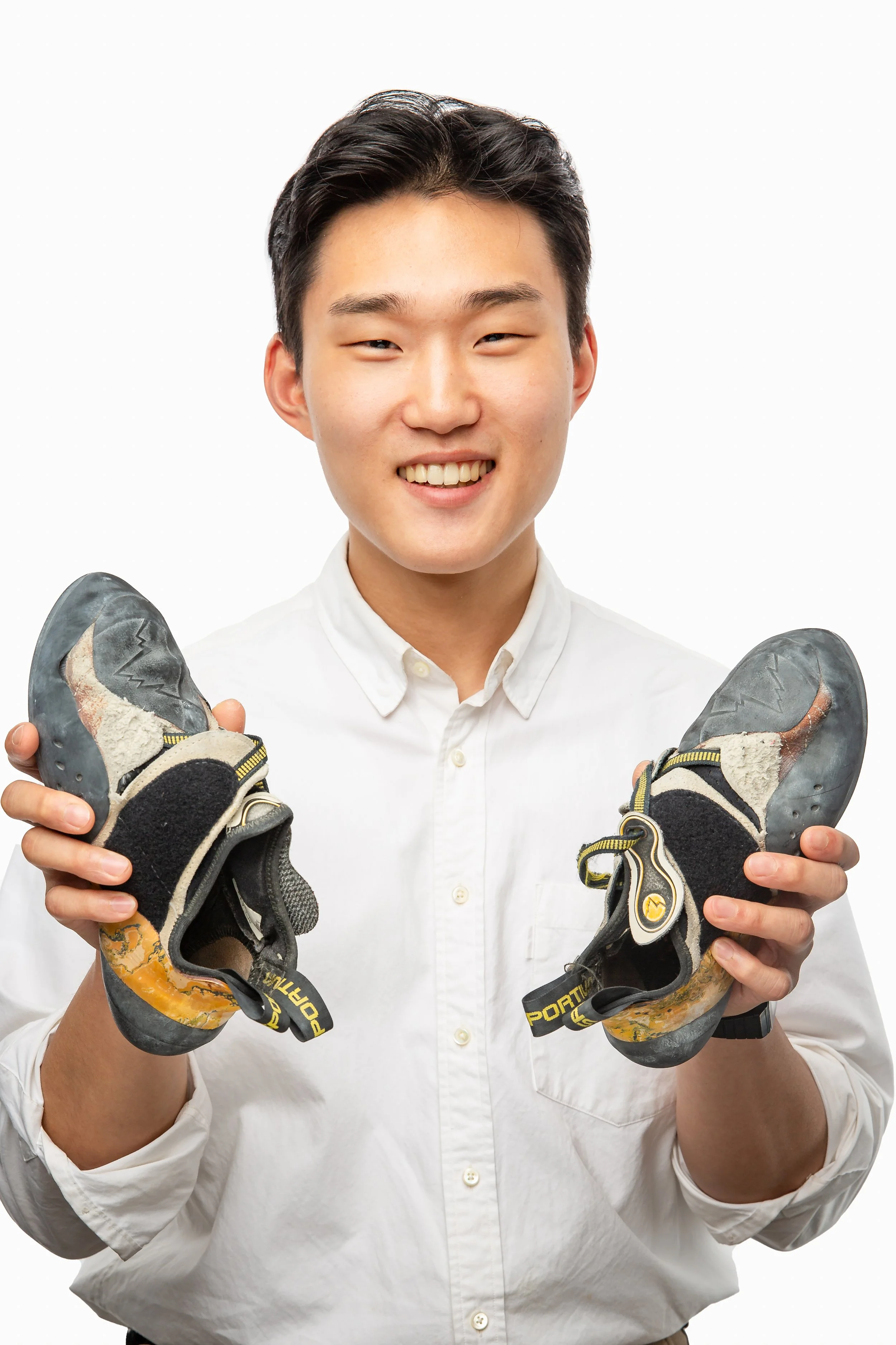 A smiling young man in a white shirt holds up a pair of worn climbing shoes, one in each hand, against a plain white background.