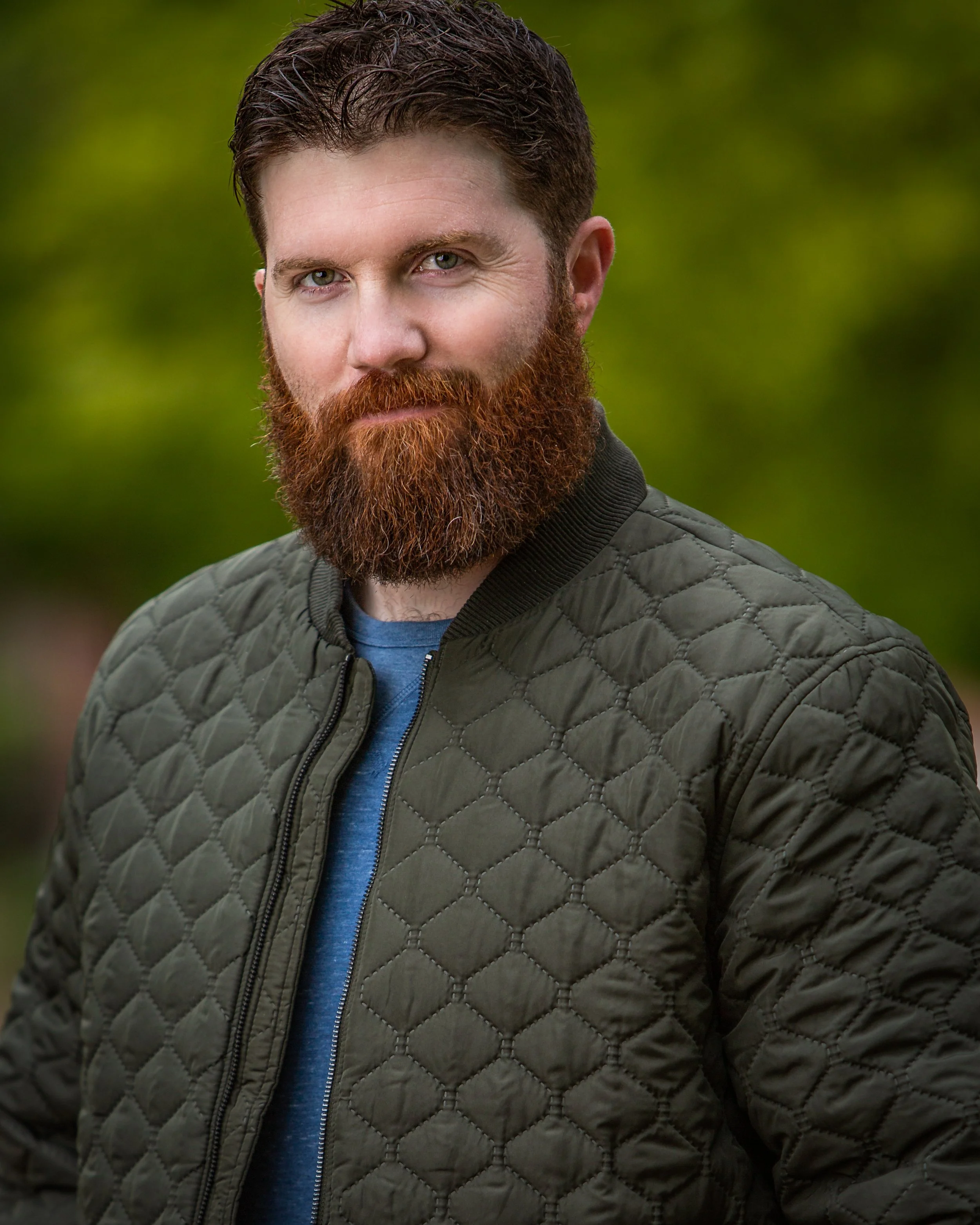 A man with a thick red beard and short brown hair wears a quilted green jacket over a blue shirt, standing outdoors with a blurred green background. He is looking at the camera with a slight smile.