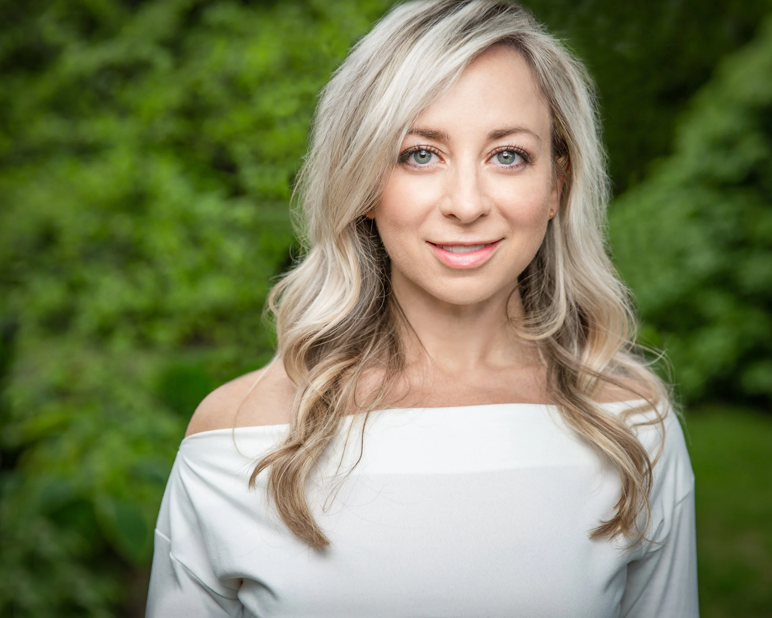 A woman with long, wavy blonde hair and blue eyes smiles softly while standing outdoors in front of lush green foliage. She is wearing an off-the-shoulder white top.