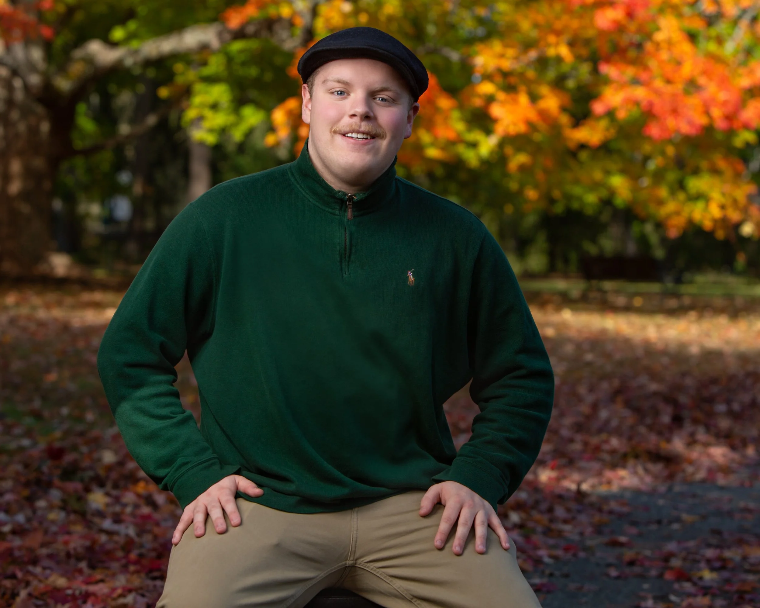 Young man smiling in a park during an outdoor senior portrait session with autumn foliage.