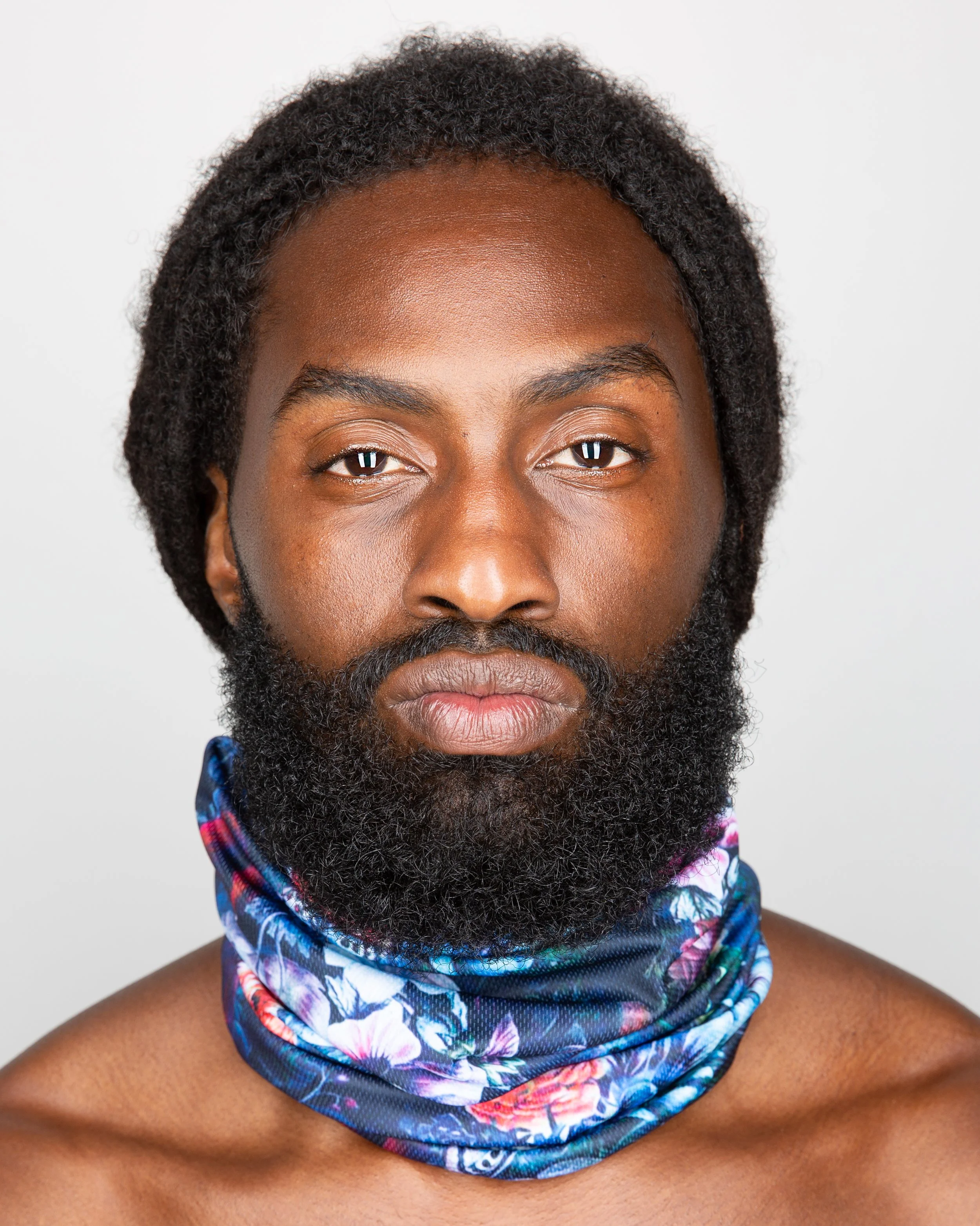 A man with a full beard and mustache wears a colorful, floral-patterned neck gaiter around his neck. He has dark hair styled in twists or locks and looks directly at the camera against a plain background.
