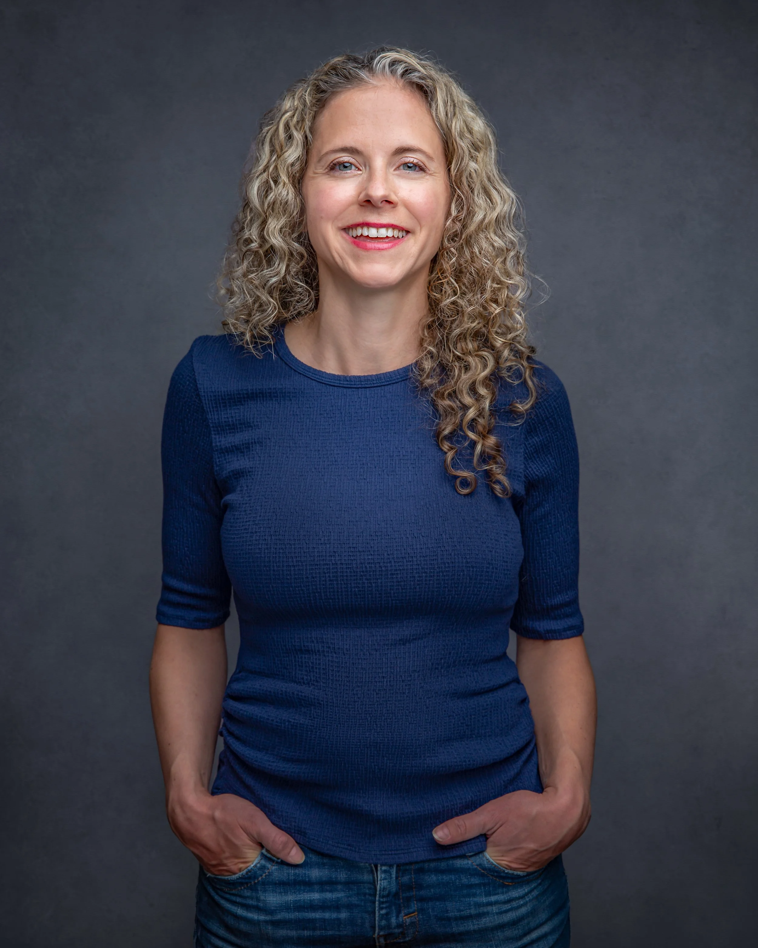 A woman with long curly blonde hair, wearing a blue shirt and jeans, stands against a dark gray background, smiling with her hands in her pockets.