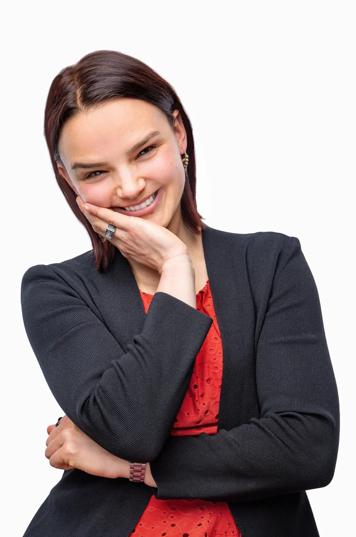 Professional headshot of a scientist with straight brown hair wearing a black blazer over a red top. She smiles warmly with her head slightly tilted, resting one hand on her cheek while her other arm is crossed against a clean white background.