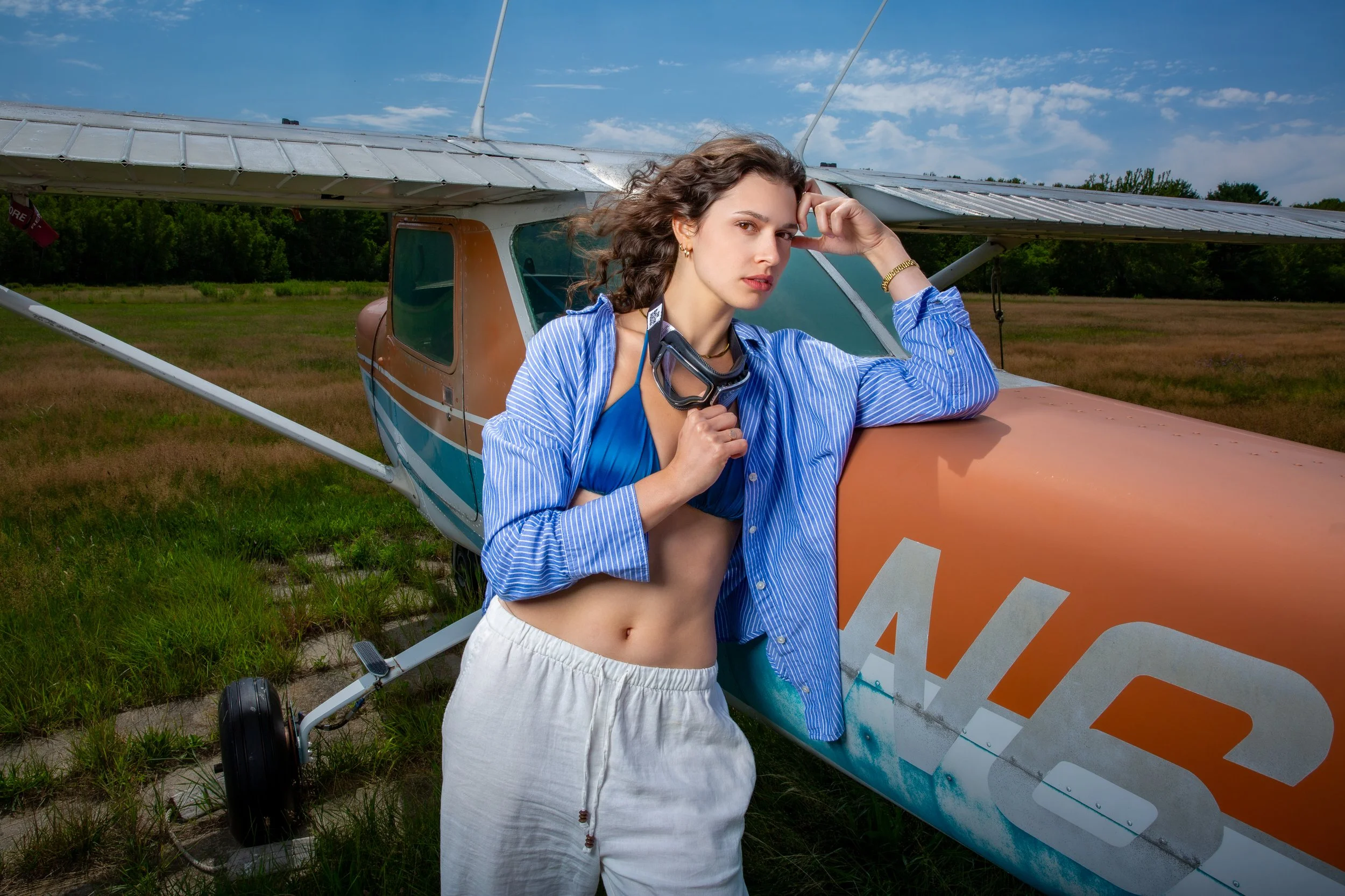 Editorial fashion portrait of a young woman posing beside a small airplane on a bright summer day.