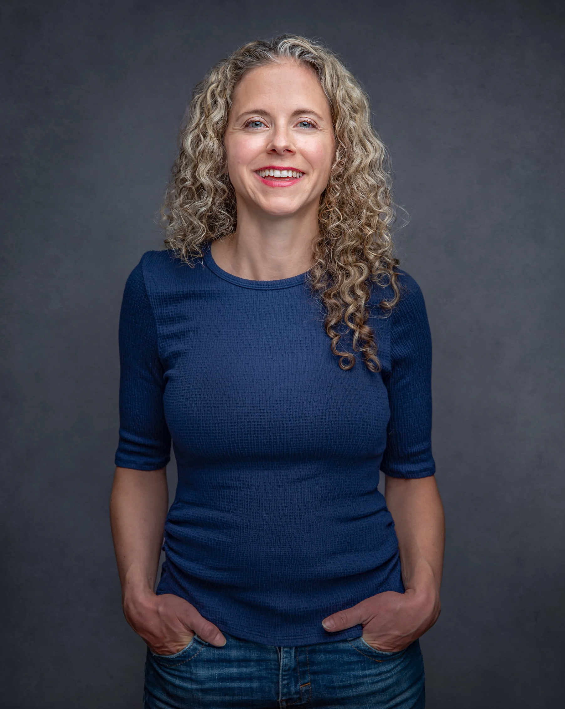 Professional headshot of an actress with long, curly blonde hair wearing a blue shirt and jeans. She stands against a dark gray background with her hands in her pockets, smiling warmly at the camera.