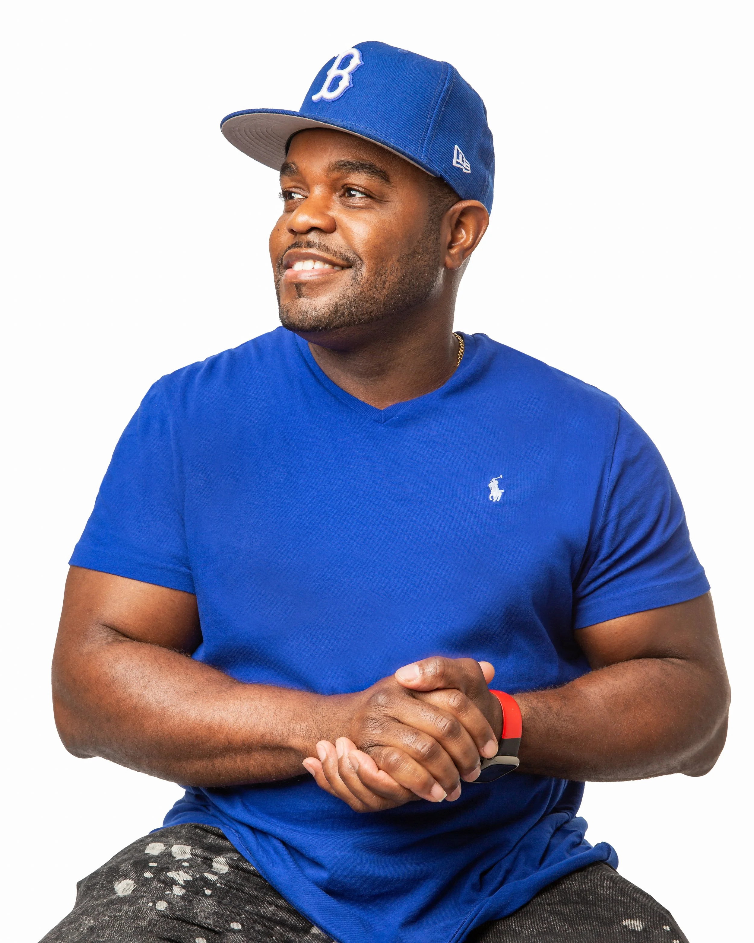 Smiling man in a blue shirt and matching blue baseball cap sits with his hands clasped, looking to the side against a white background.