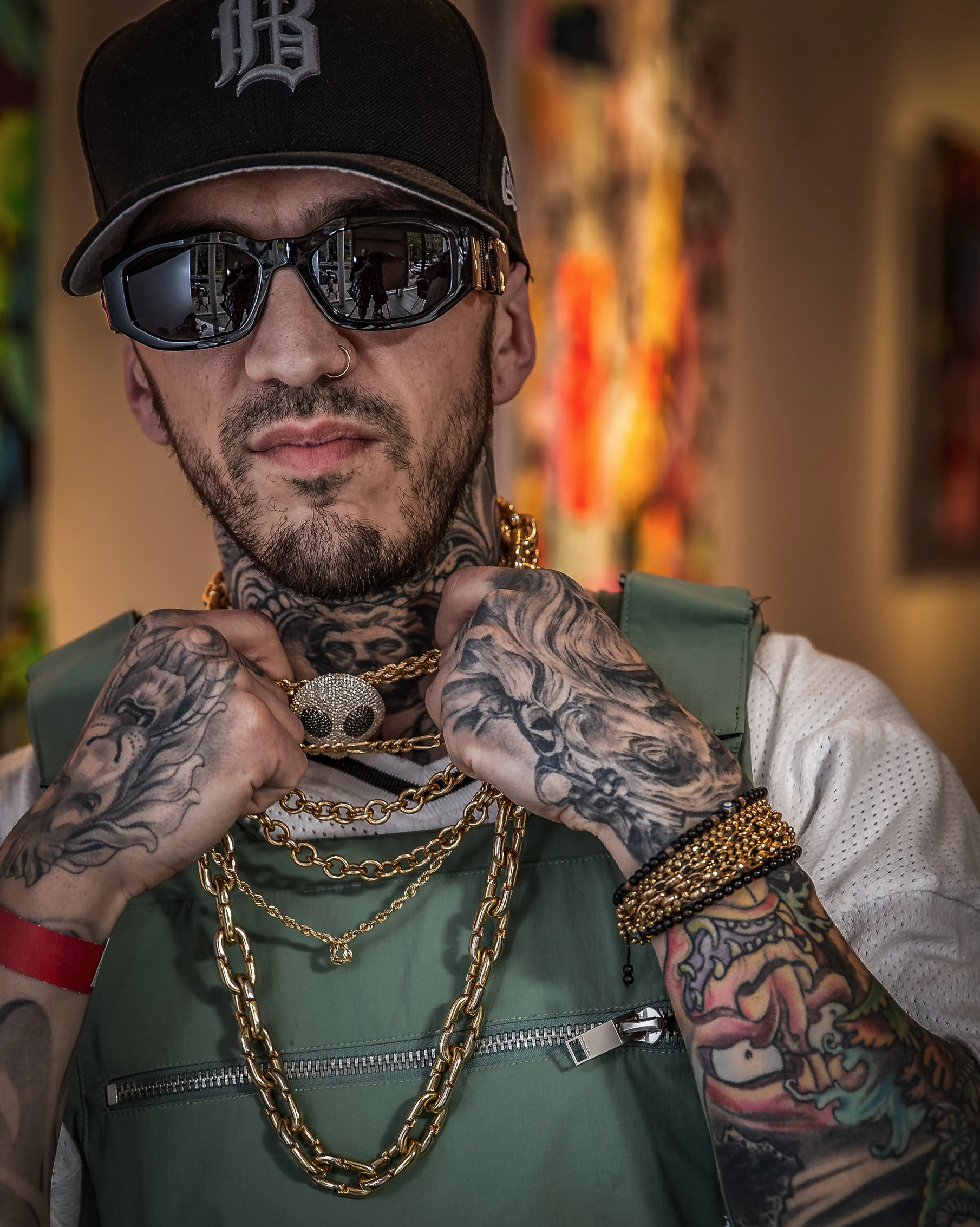 Artist portrait of rapper Timmy Sneaks wearing a black cap, sunglasses, a green vest over a white shirt, and layered gold chains. His tattooed hands and distinctive style make for a bold contemporary portrait.