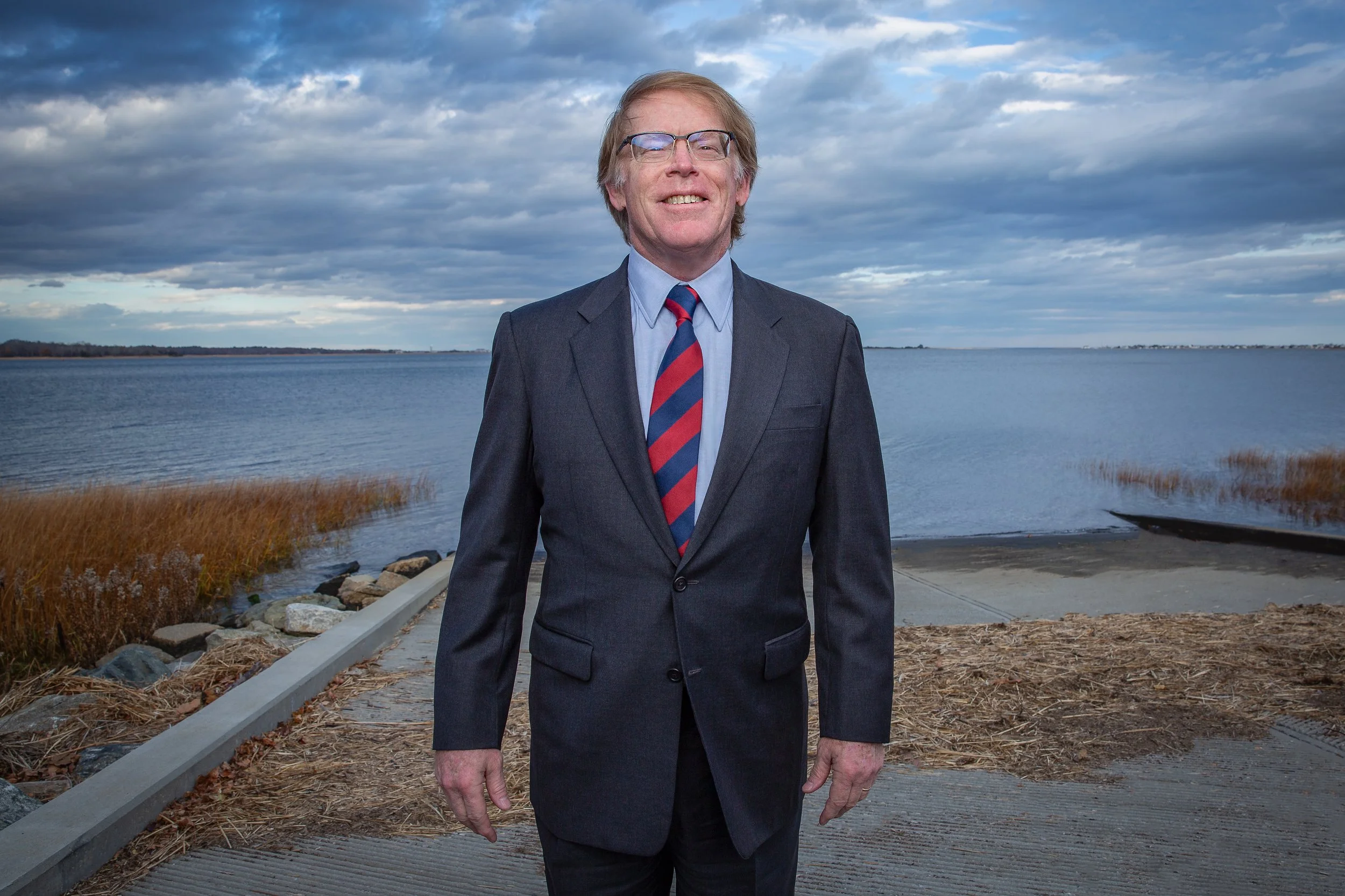 A man in a dark suit, blue shirt, and red-and-blue striped tie stands smiling near a lake or river with cloudy skies and reeds in the background.