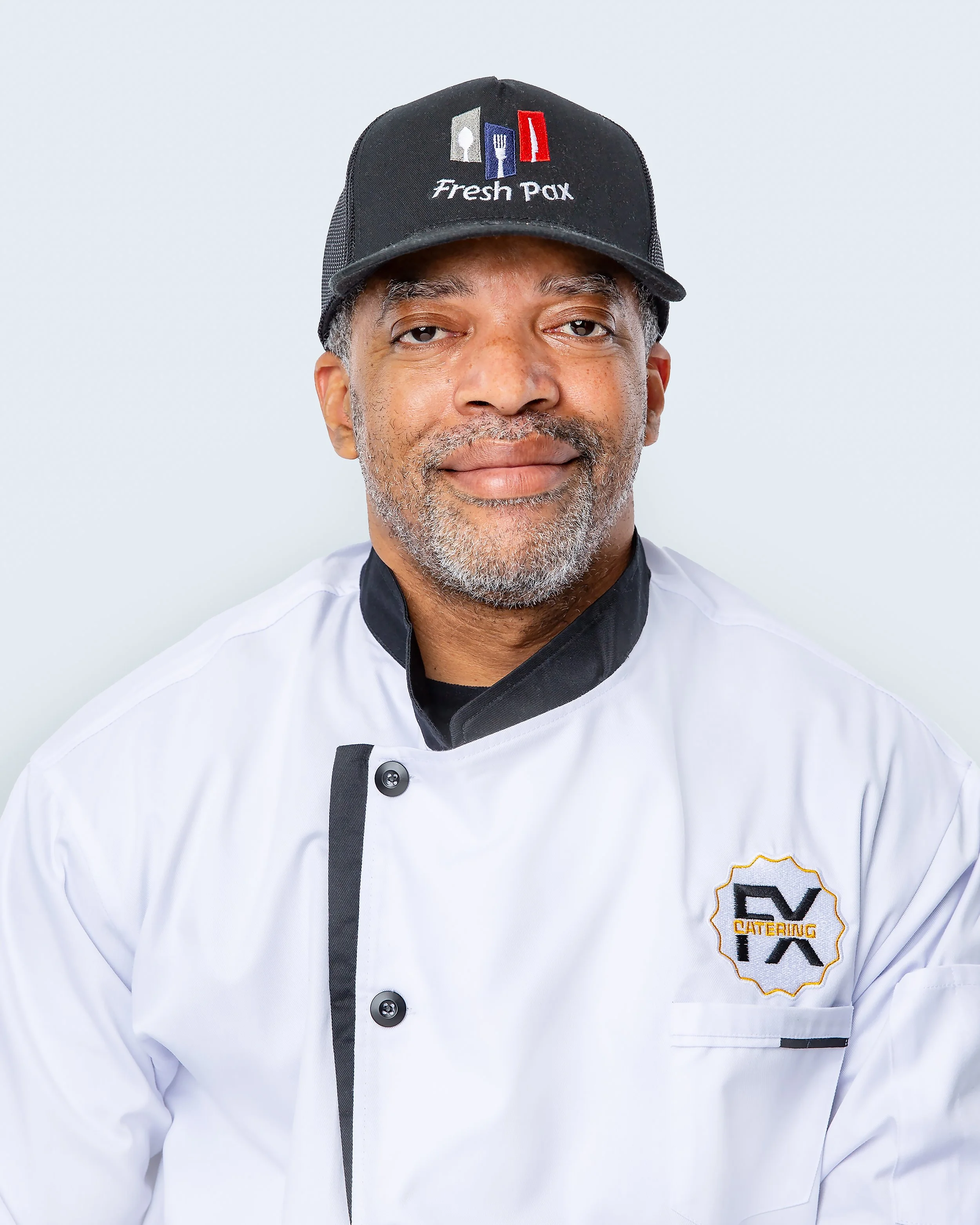 Business headshot of a chef wearing a white chef's coat and a black Fresh Pax cap. He looks directly at the camera with a warm, confident expression against a clean light gray background.