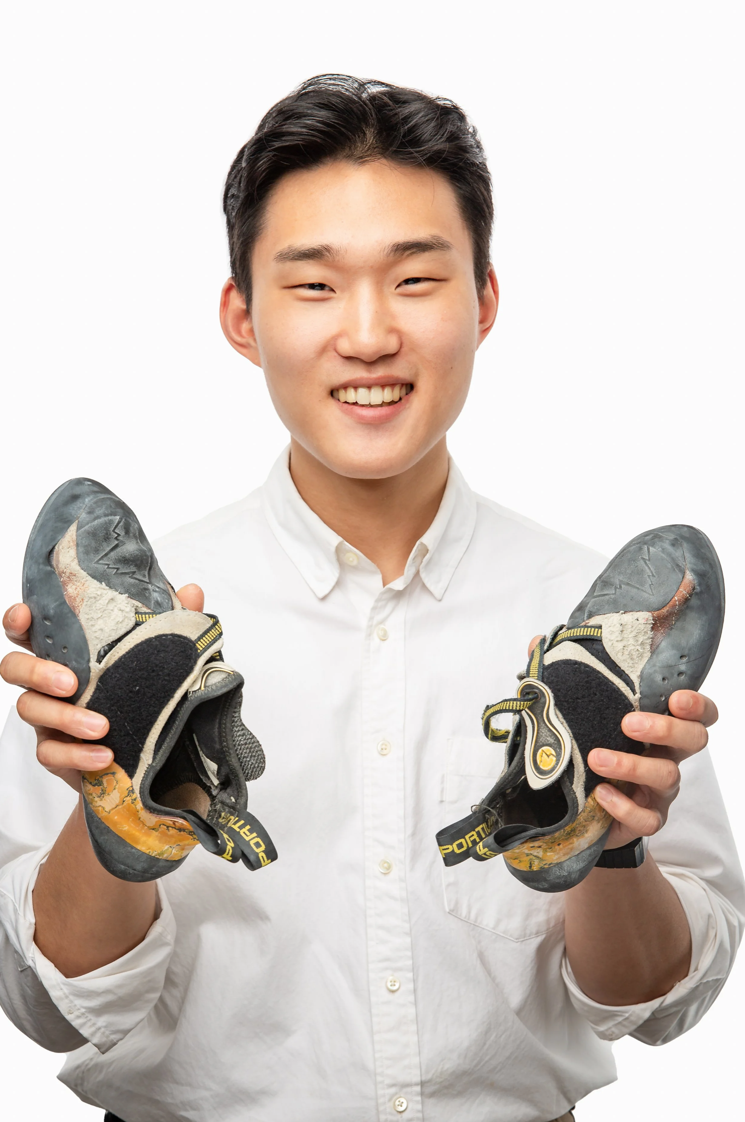 Environmental headshot of a smiling young man wearing a white shirt and holding a pair of well-worn bike riding shoes. He stands against a clean white background, highlighting his passion for cycling.