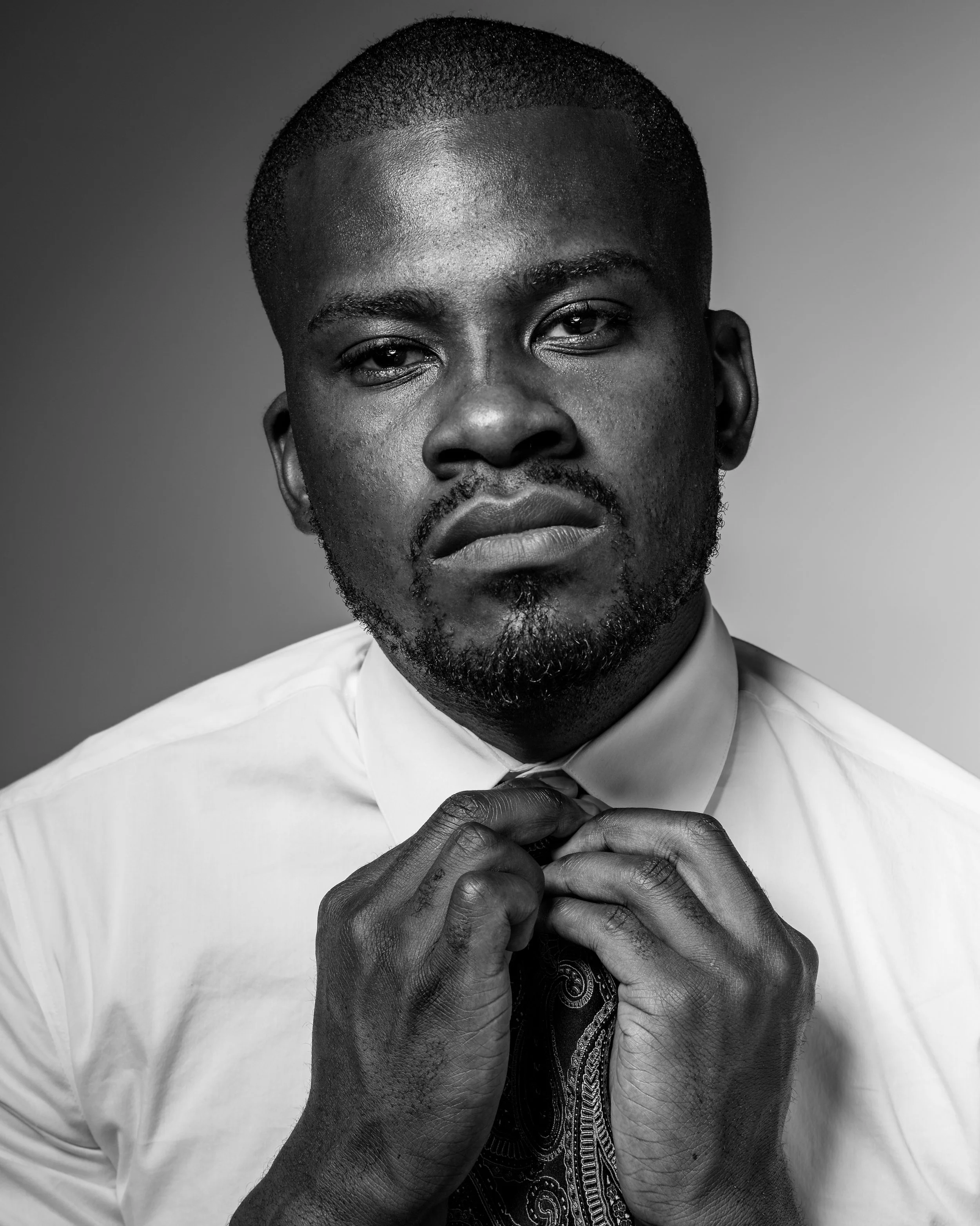 Black and white portrait of a man in a white shirt adjusting his tie, looking seriously at the camera with a neutral background.