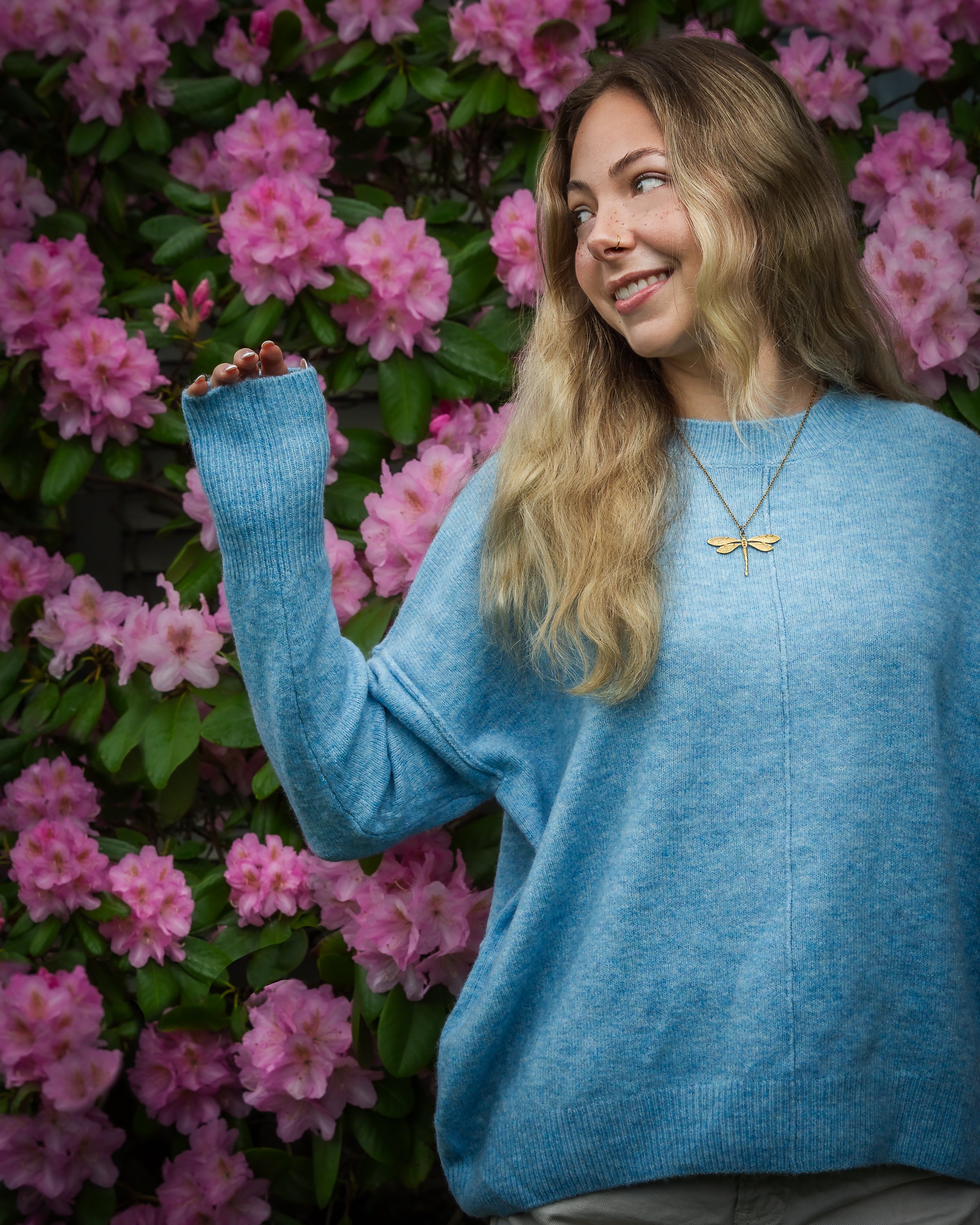 High school senior smiling beside blooming pink rhododendrons during an outdoor portrait session.