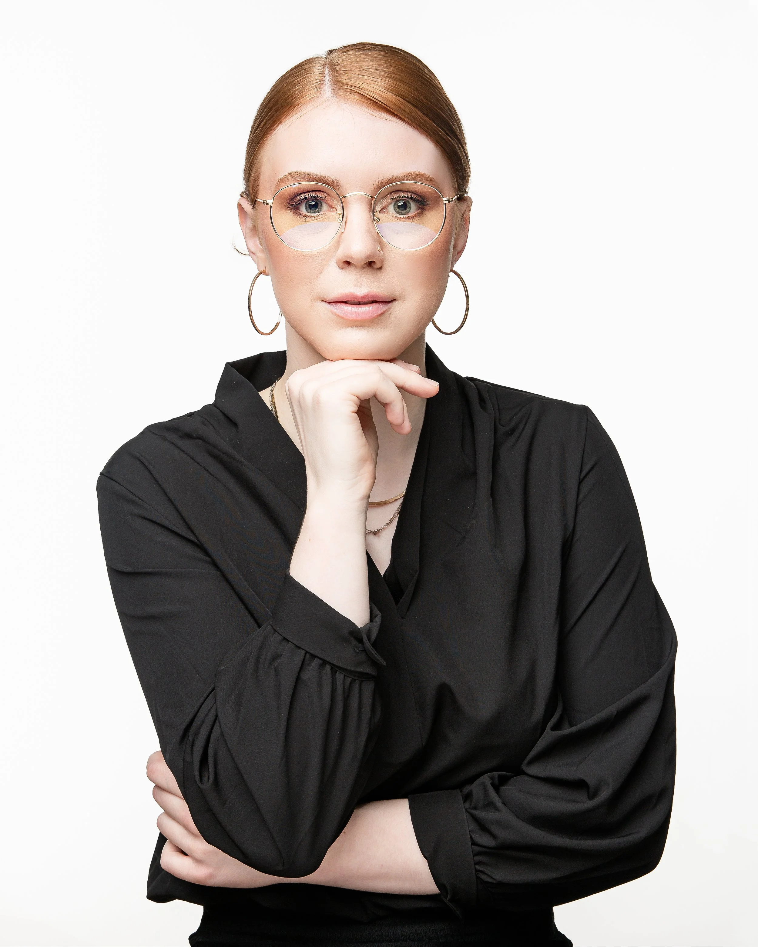 Studio headshot of a woman with red hair wearing round glasses and a black blouse. She looks directly at the camera with a calm, confident expression, resting her chin on her hand against a clean white background.