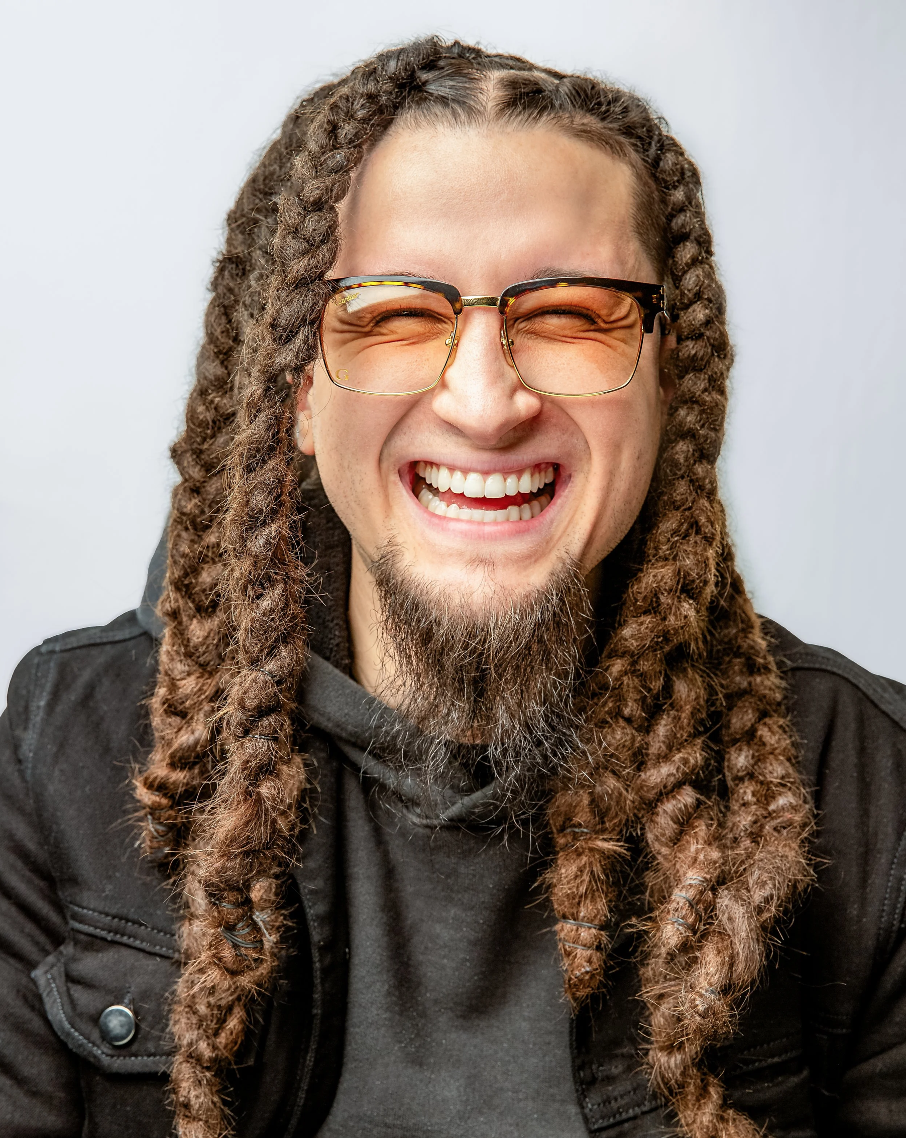 Lifestyle portrait of rapper Yo-K with long, thick braids, a goatee, and tinted glasses. He smiles broadly while wearing a black jacket over a hoodie against a clean, light-colored background.