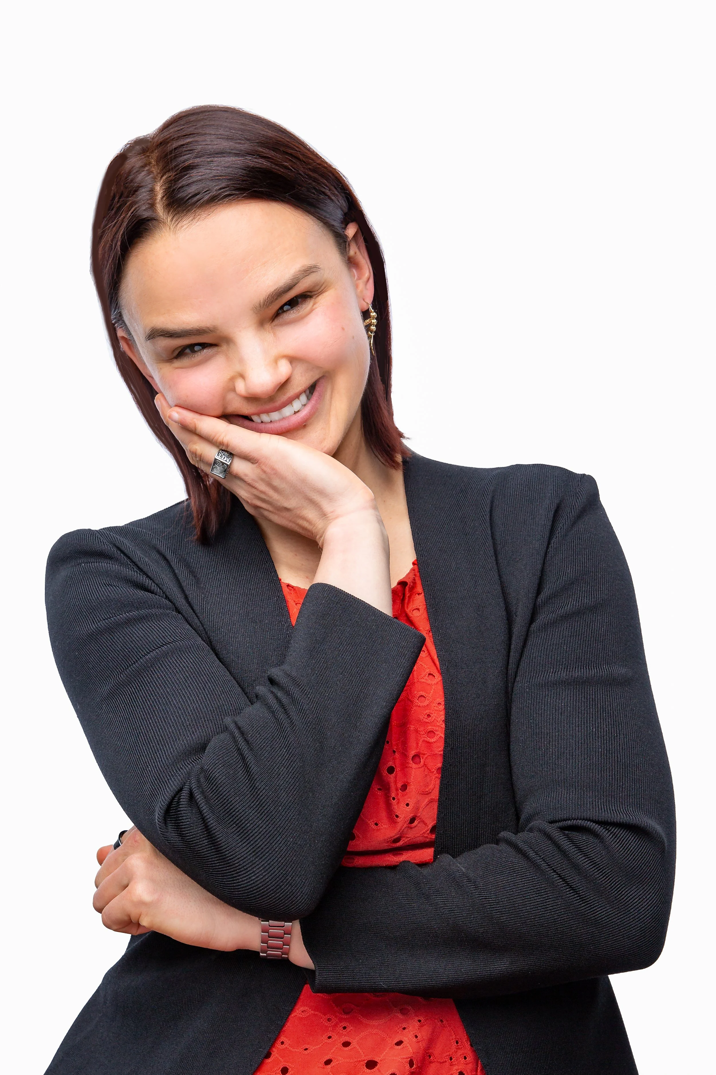 A woman with straight brown hair, wearing a black blazer over a red top, smiles with her head tilted and her hand resting on her cheek. She has her other arm crossed and stands against a white background.