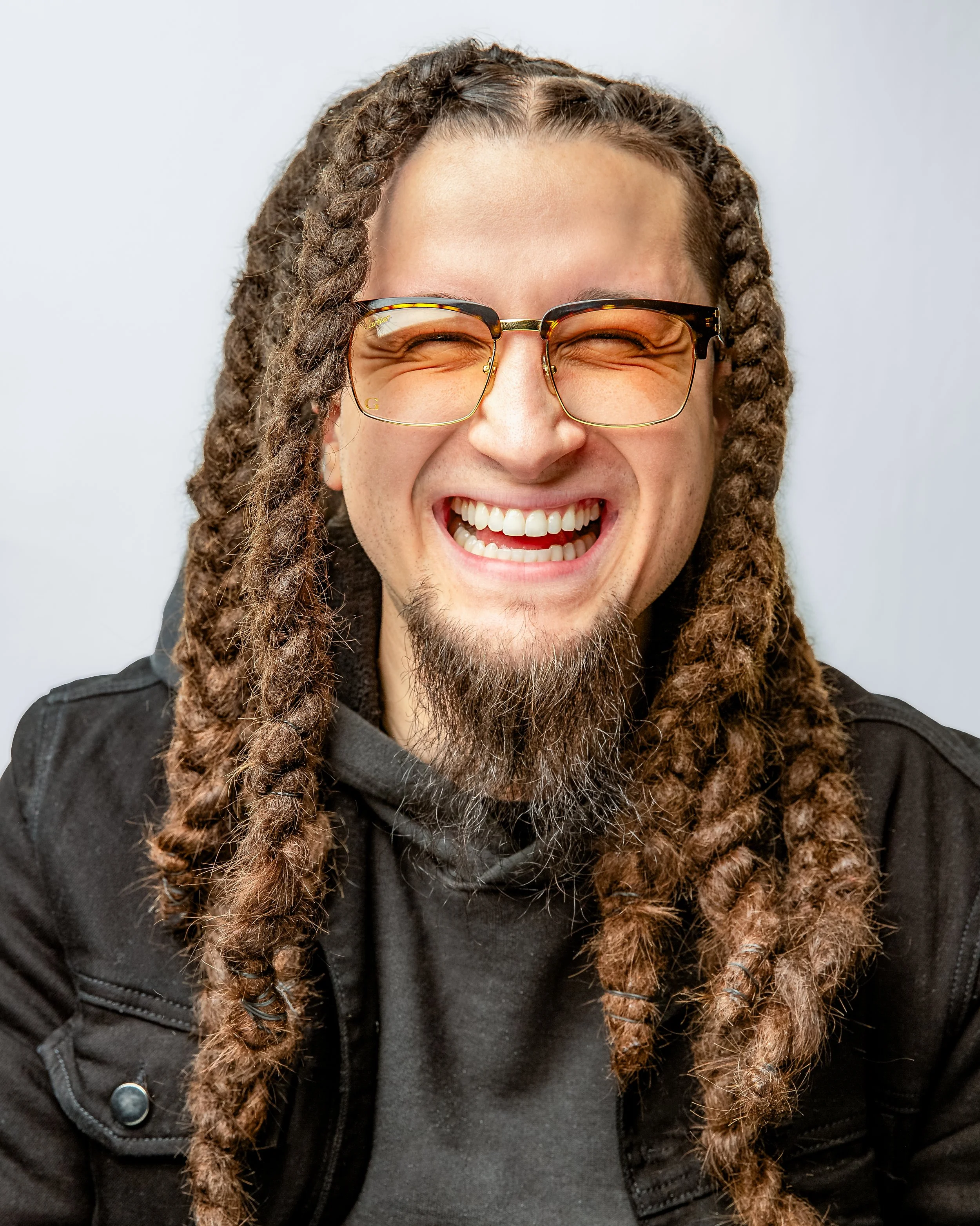 A man with long, thick braids, a goatee, and mustache smiles widely while wearing tinted glasses and a black jacket over a hoodie. The background is plain and light-colored.