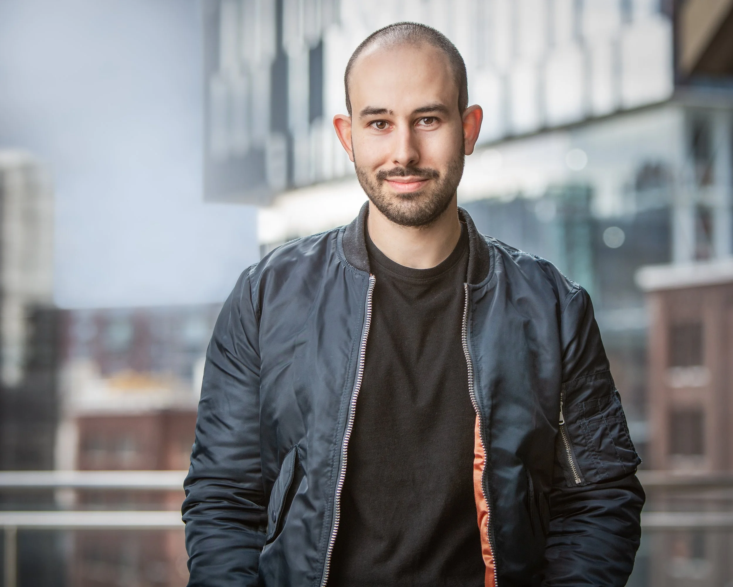 A man with a shaved head and beard, wearing a black bomber jacket and black shirt, stands outside in an urban setting with modern buildings in the blurred background. He is smiling slightly and facing the camera.