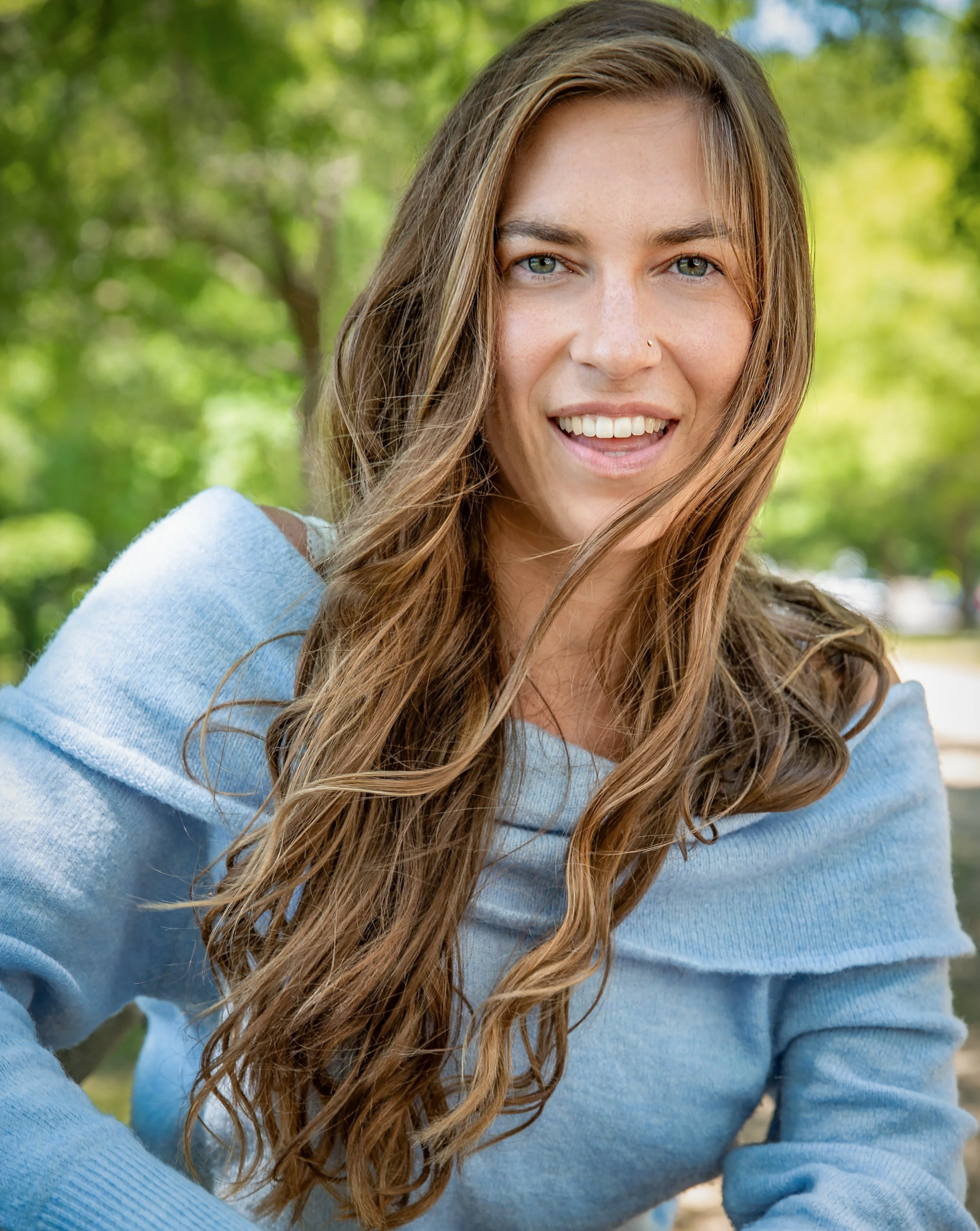Outdoor portrait of a smiling woman with long, wavy brown hair and blue eyes wearing a light blue off-the-shoulder sweater. She is seated in front of softly blurred green trees, creating a warm and approachable feel.