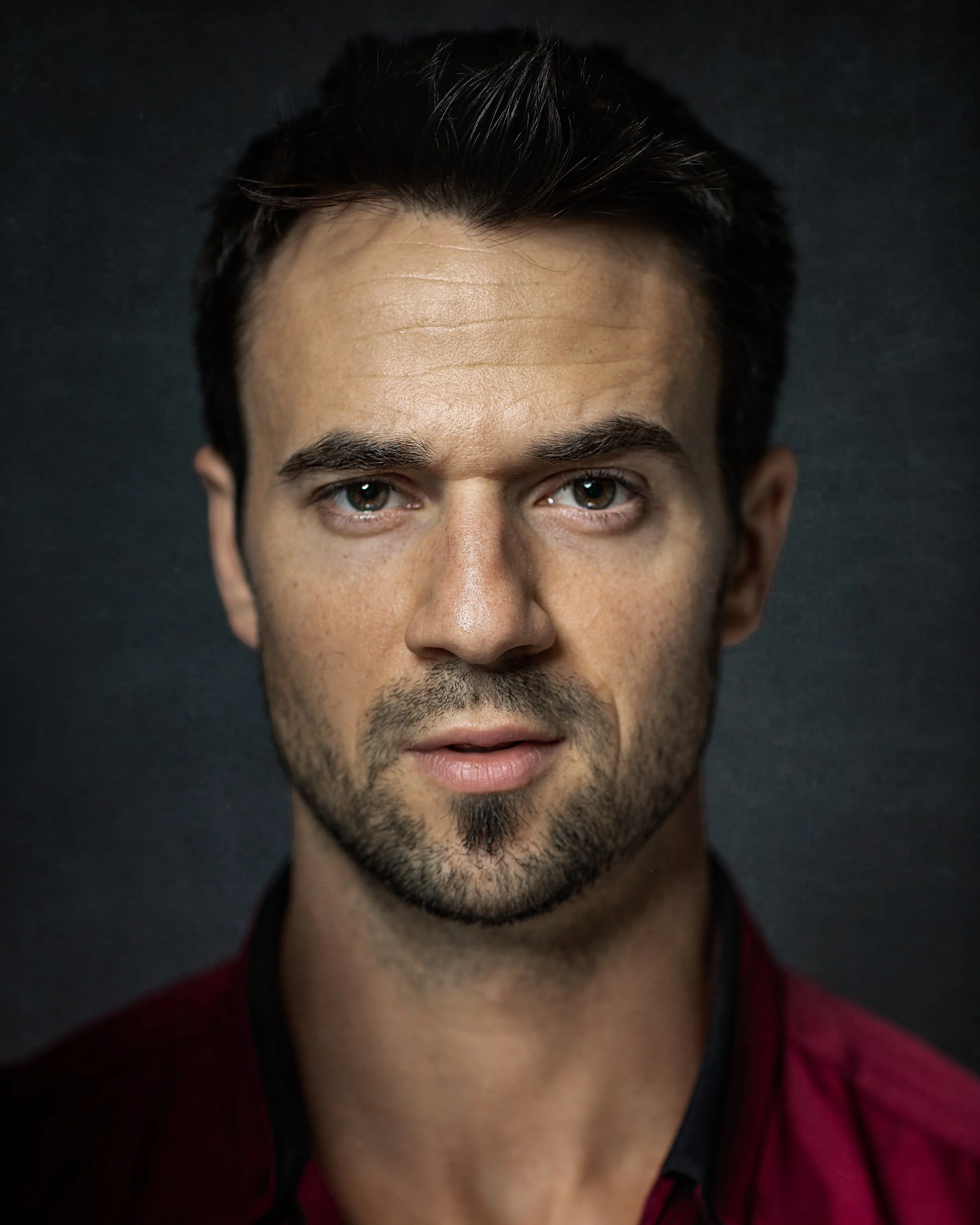 A man with short dark hair and facial stubble looks directly at the camera with a neutral expression. He is wearing a dark red shirt and is photographed against a dark, plain background.