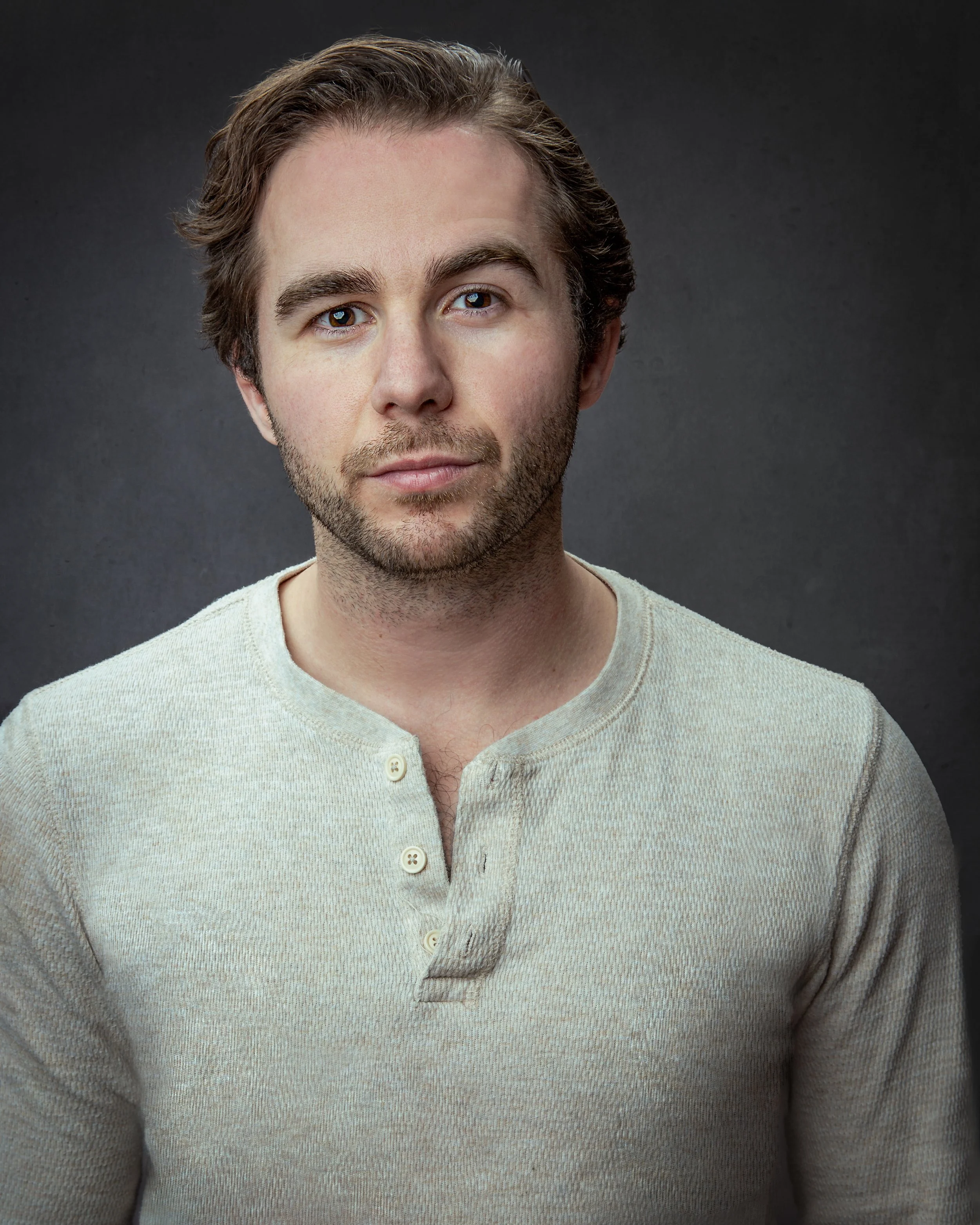 A man with light brown hair and a short beard is wearing a light-colored, long-sleeve henley shirt and looking directly at the camera with a neutral expression against a dark gray background.