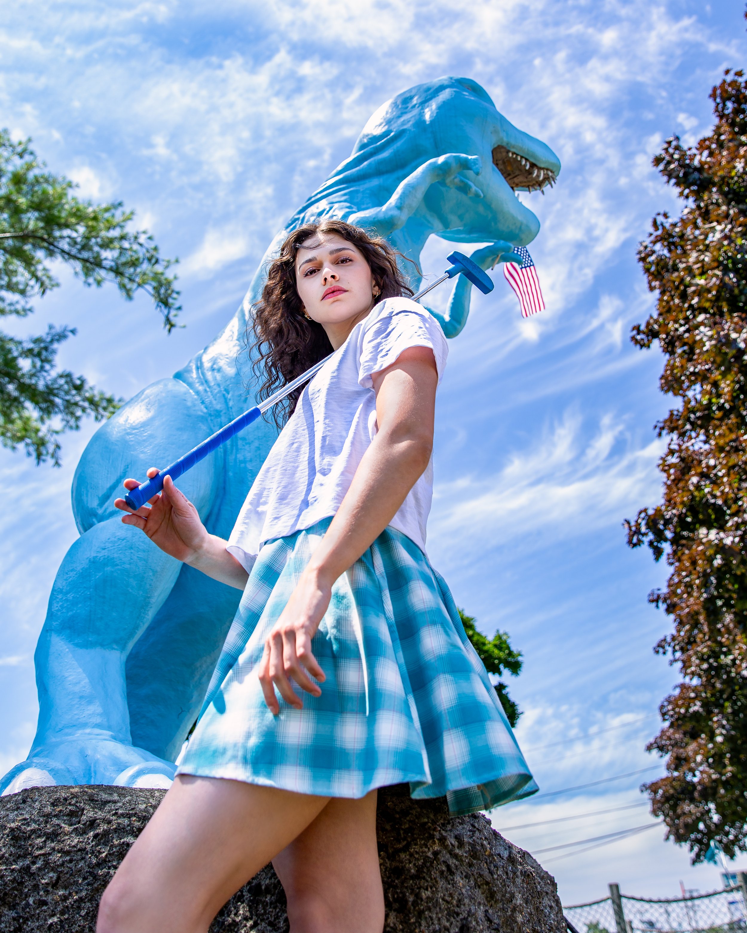 Editorial portrait of a young woman holding a golf club beneath a large blue dinosaur sculpture at an outdoor mini golf course.