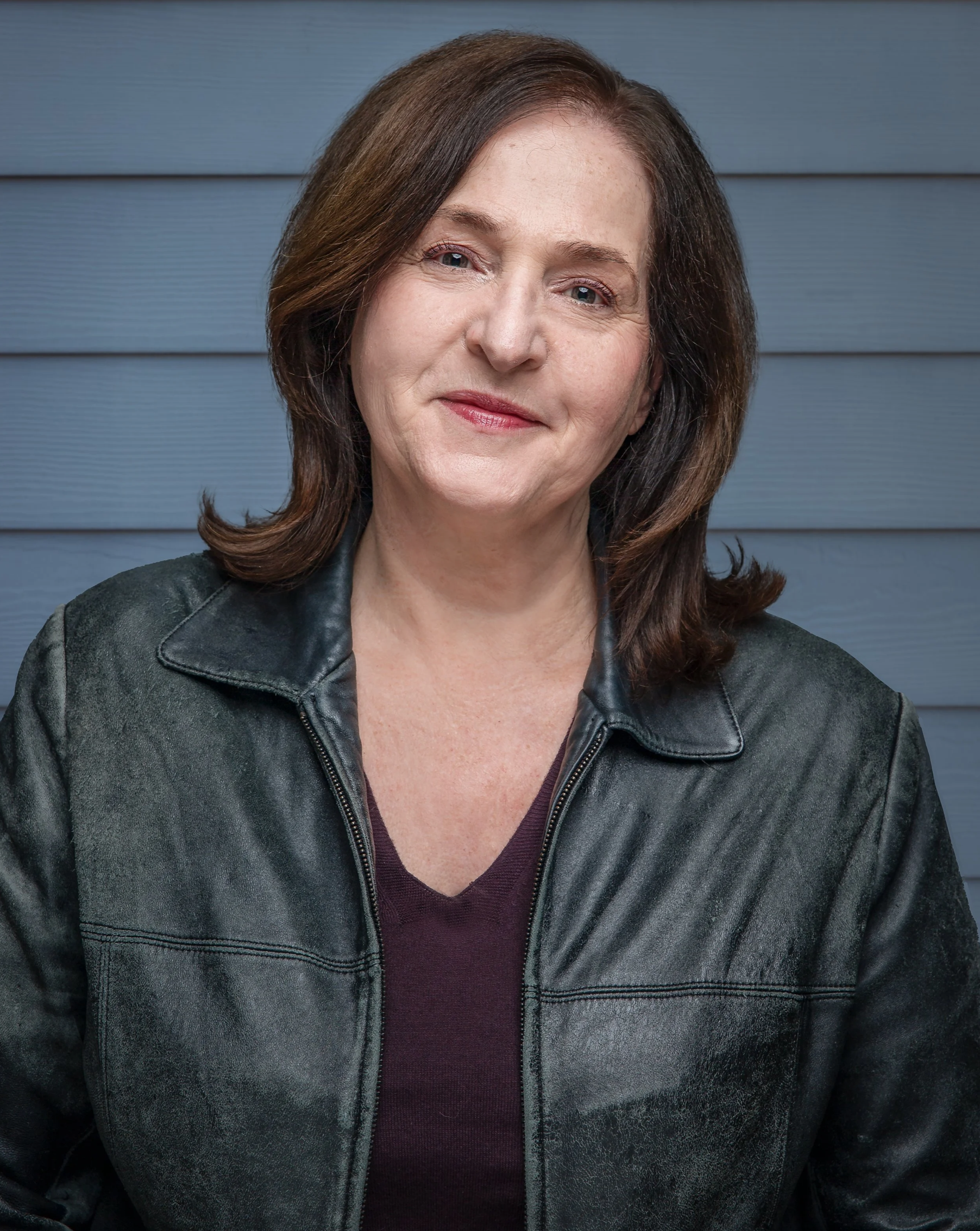 Environmental portrait of playwright Deirdre Girard wearing a dark leather jacket over a burgundy top. She smiles warmly while standing in front of a blue paneled wall, creating a relaxed and approachable portrait.