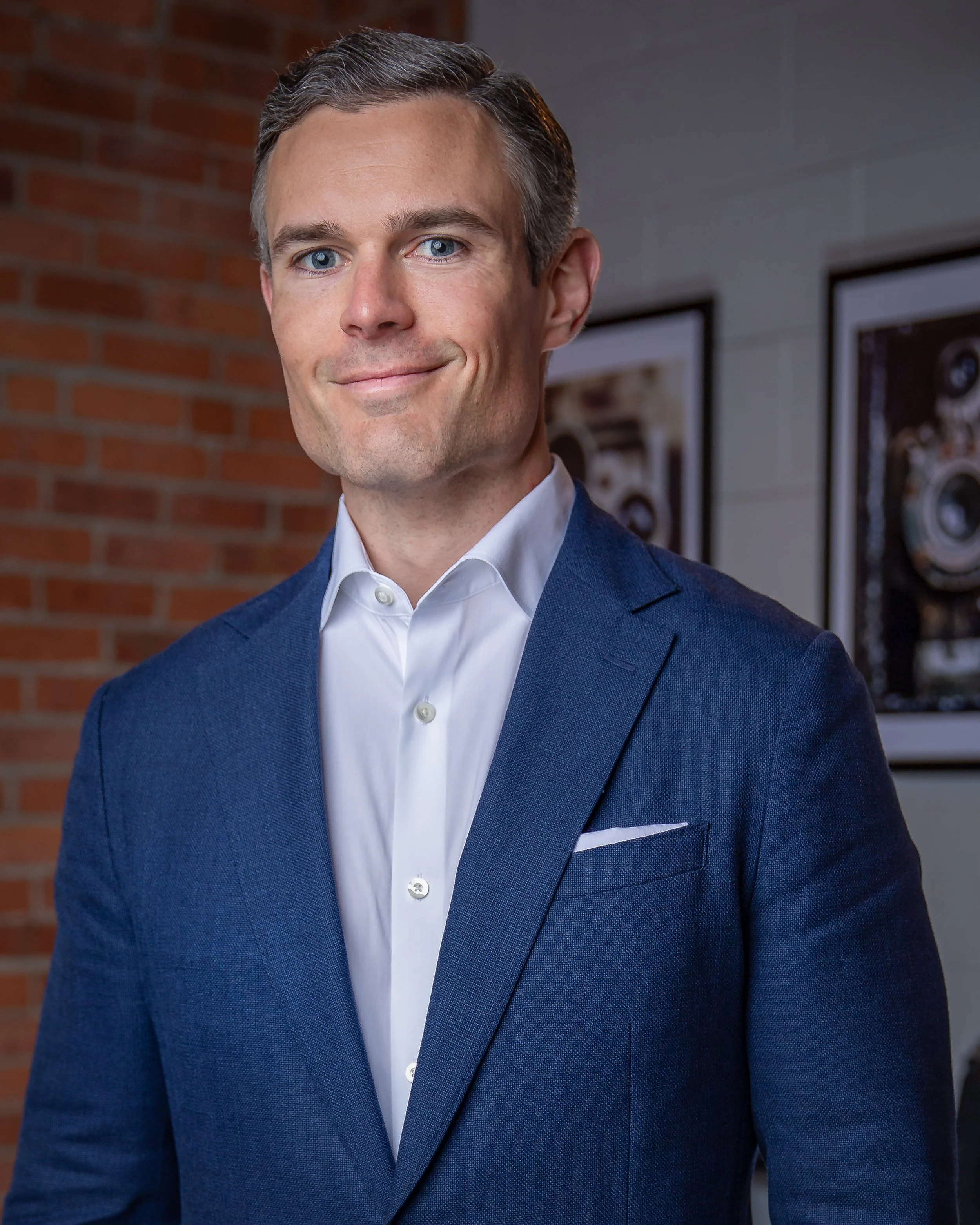 A man in a blue suit and white dress shirt stands indoors, smiling slightly. He is in front of a brick wall and framed photos of vintage cameras.