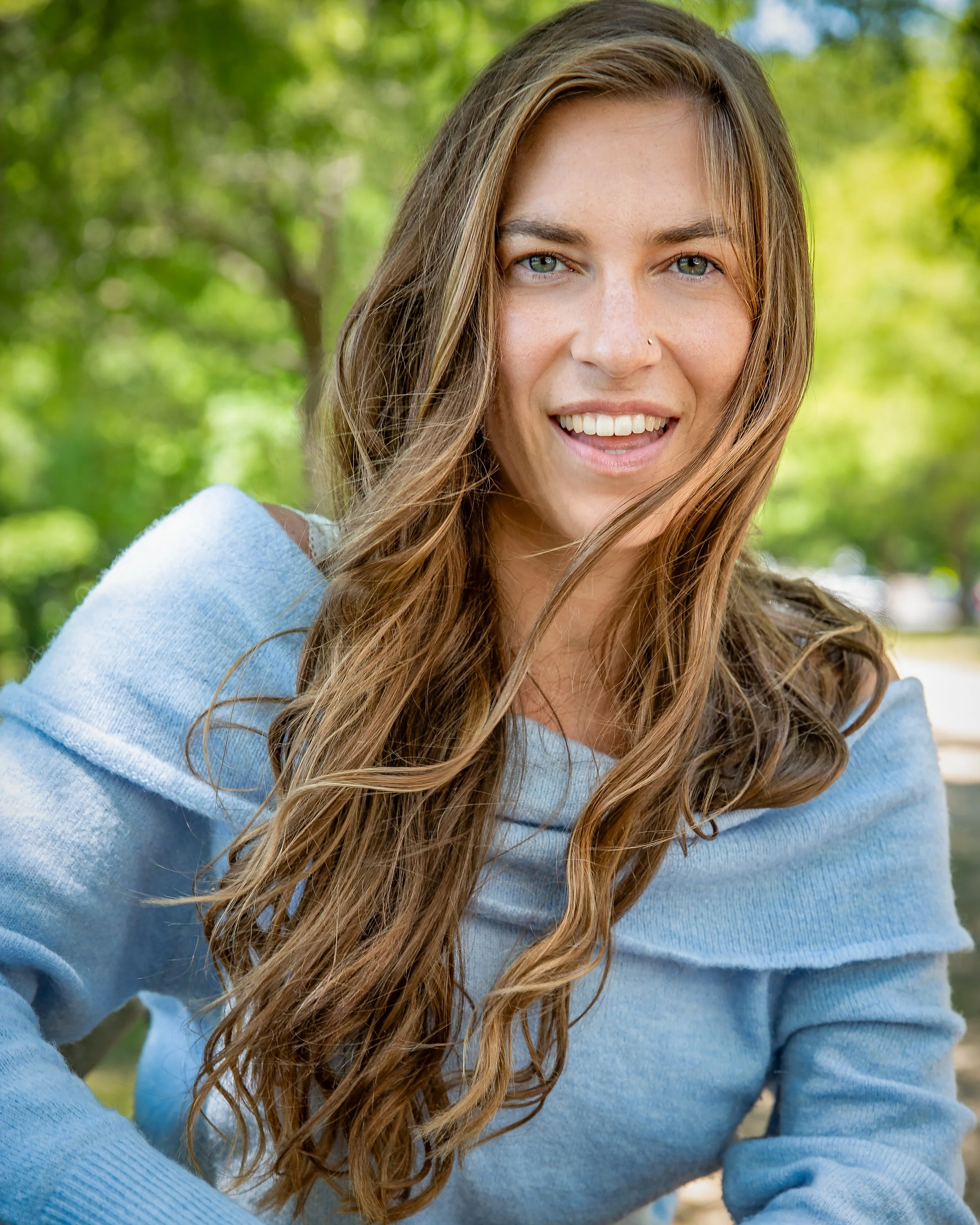 A smiling woman with long, wavy brown hair and blue eyes wears a light blue off-the-shoulder sweater, sitting outdoors with green trees in the background.