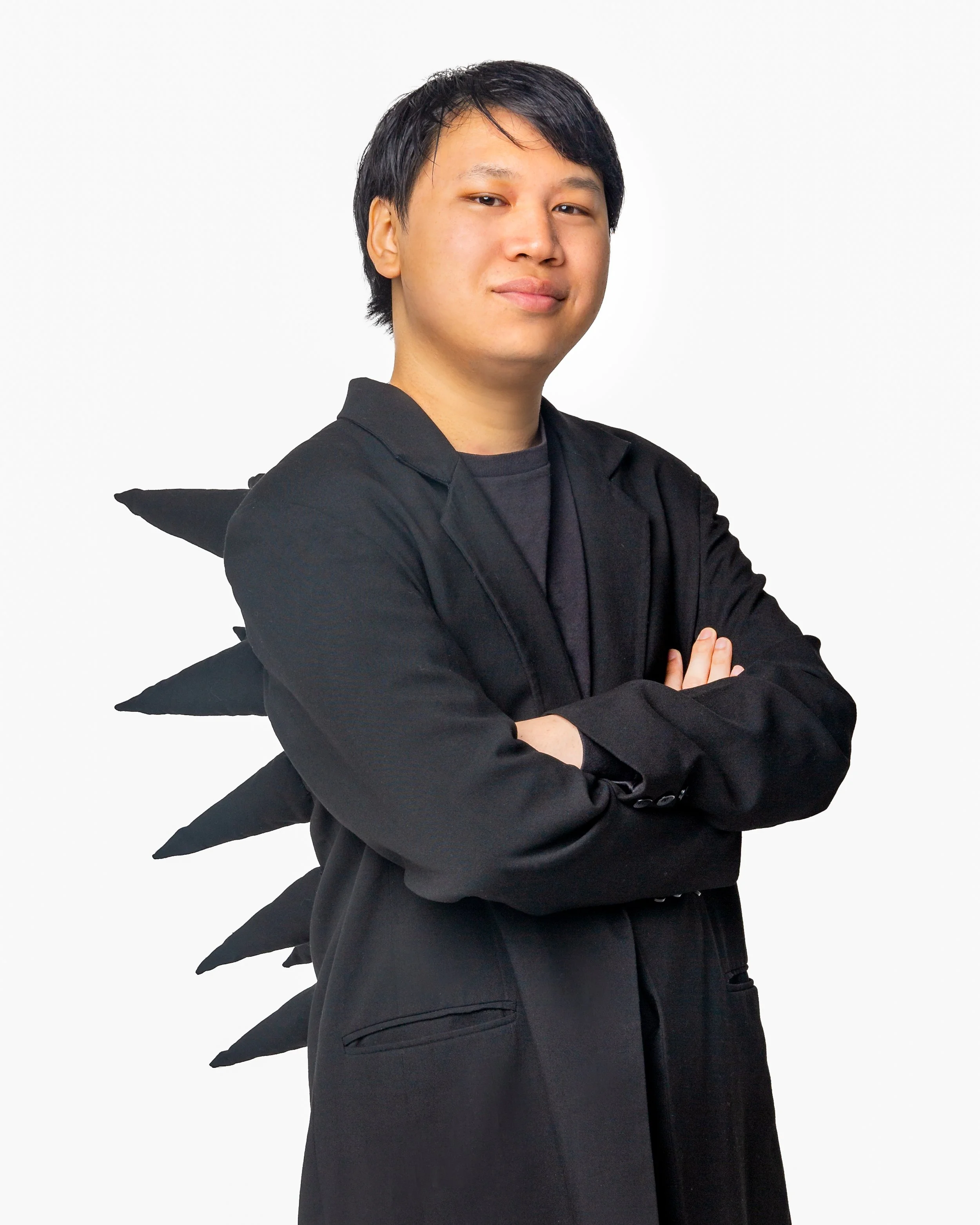 A person with black hair, wearing a black outfit and a jacket with black spikes on the back, stands confidently with arms crossed against a plain white background.