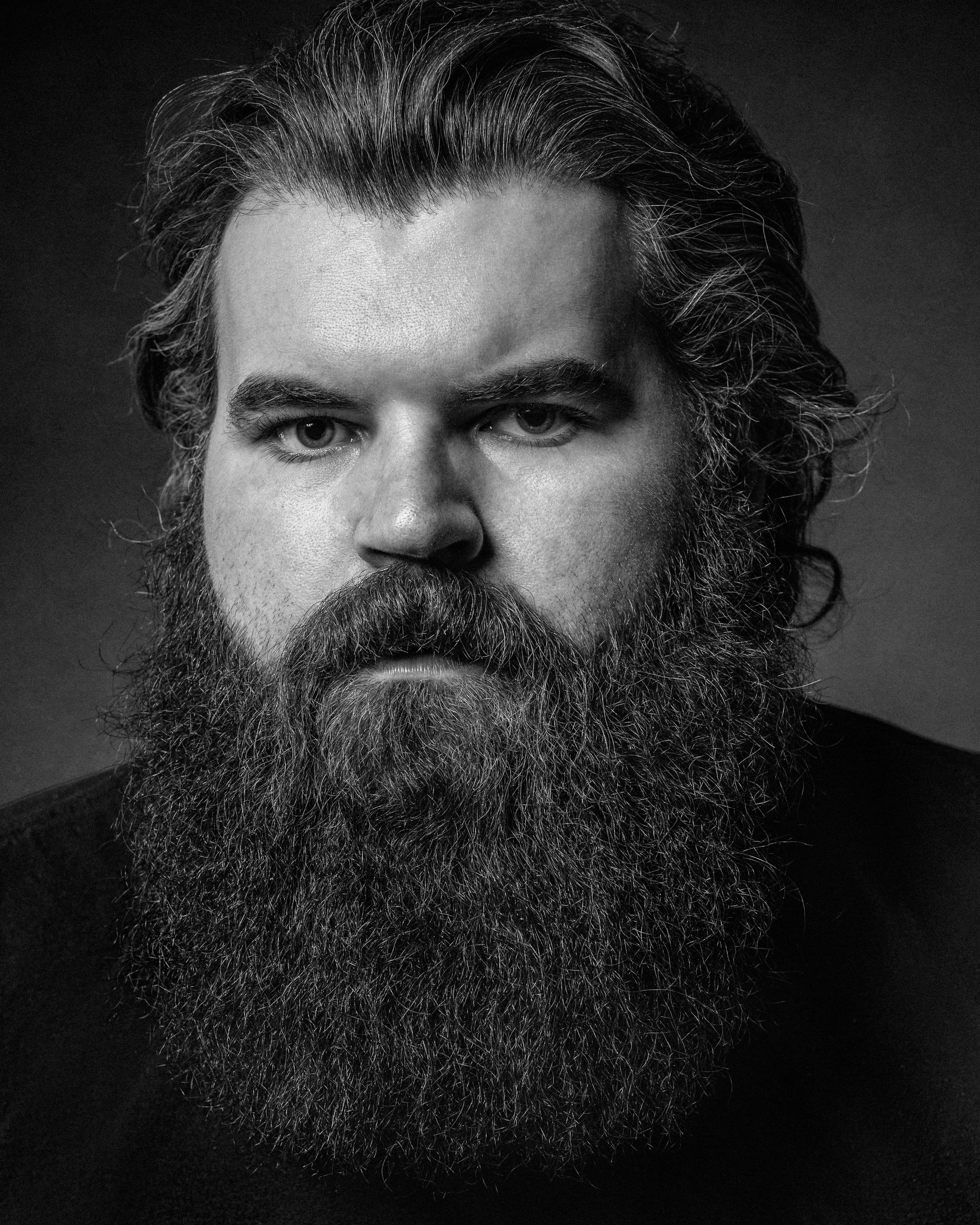 Black and white portrait of a man with thick, wavy hair and a full, bushy beard. He has a serious expression and is wearing a dark shirt against a dark background.