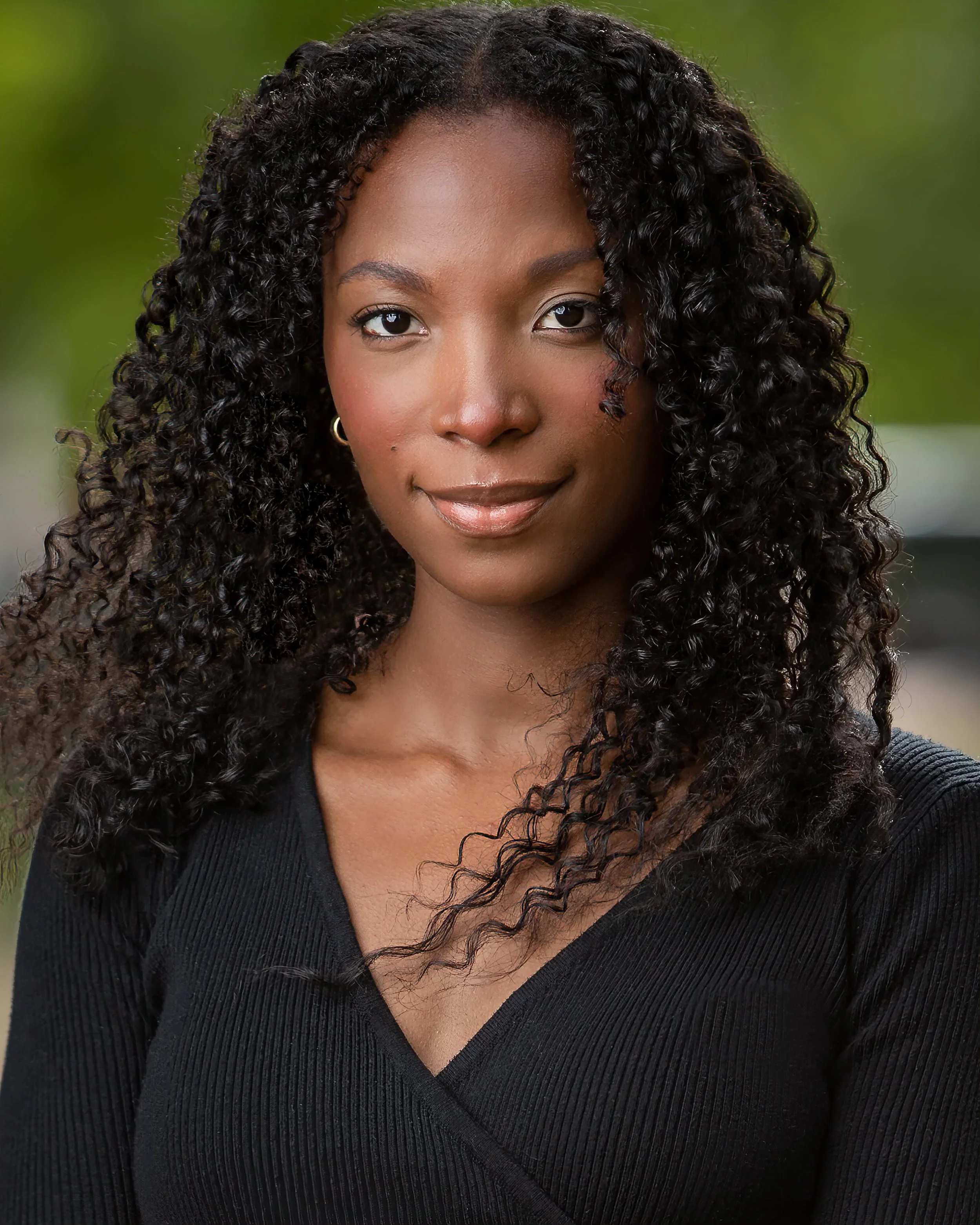 A woman with curly black hair wearing a black V-neck top stands outdoors, looking confidently at the camera with a slight smile. The background is blurred and green.