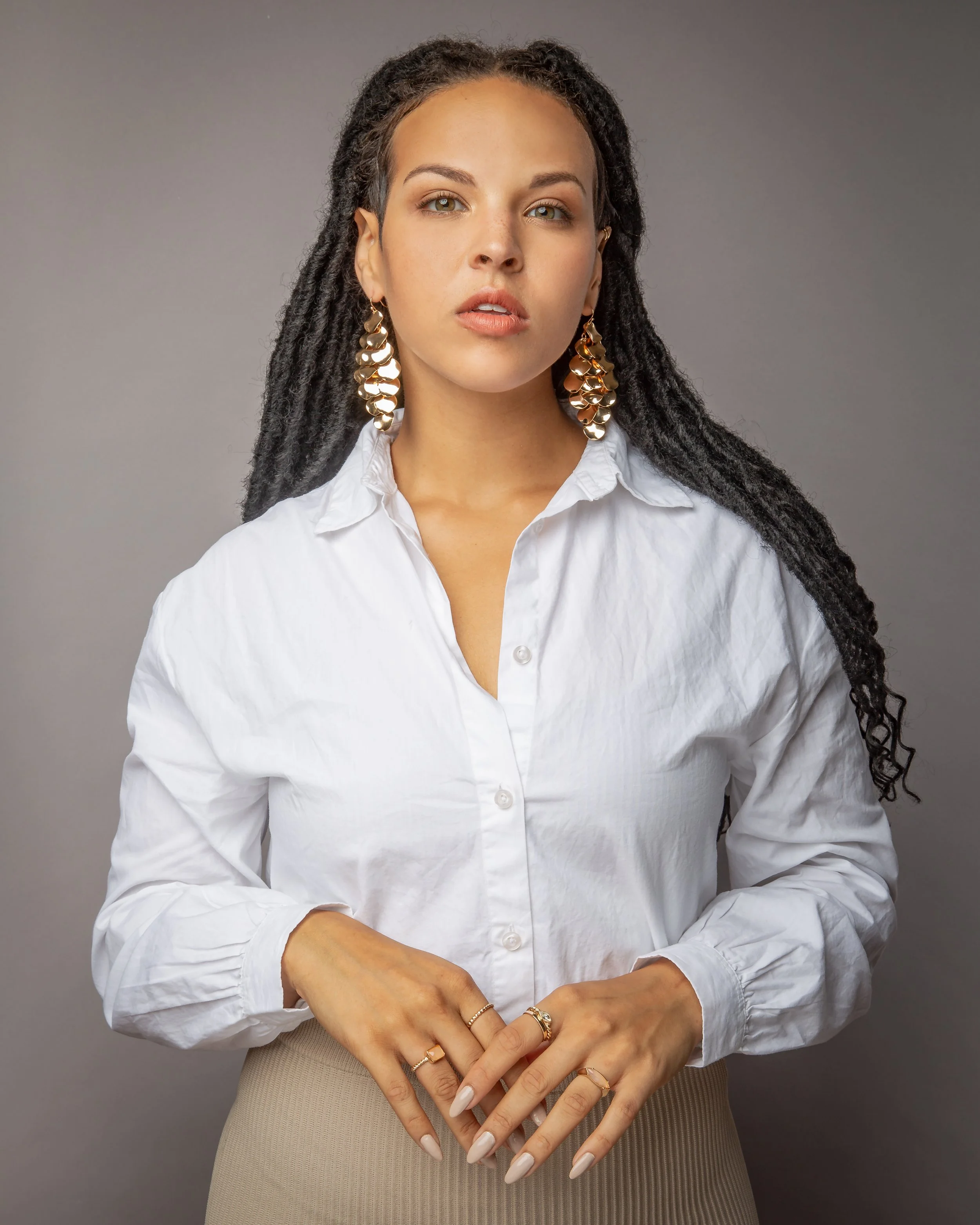 A woman with long, dark, braided hair wears a white button-up shirt, gold statement earrings, and rings. She stands against a plain gray background, looking confidently at the camera with her hands gently clasped.