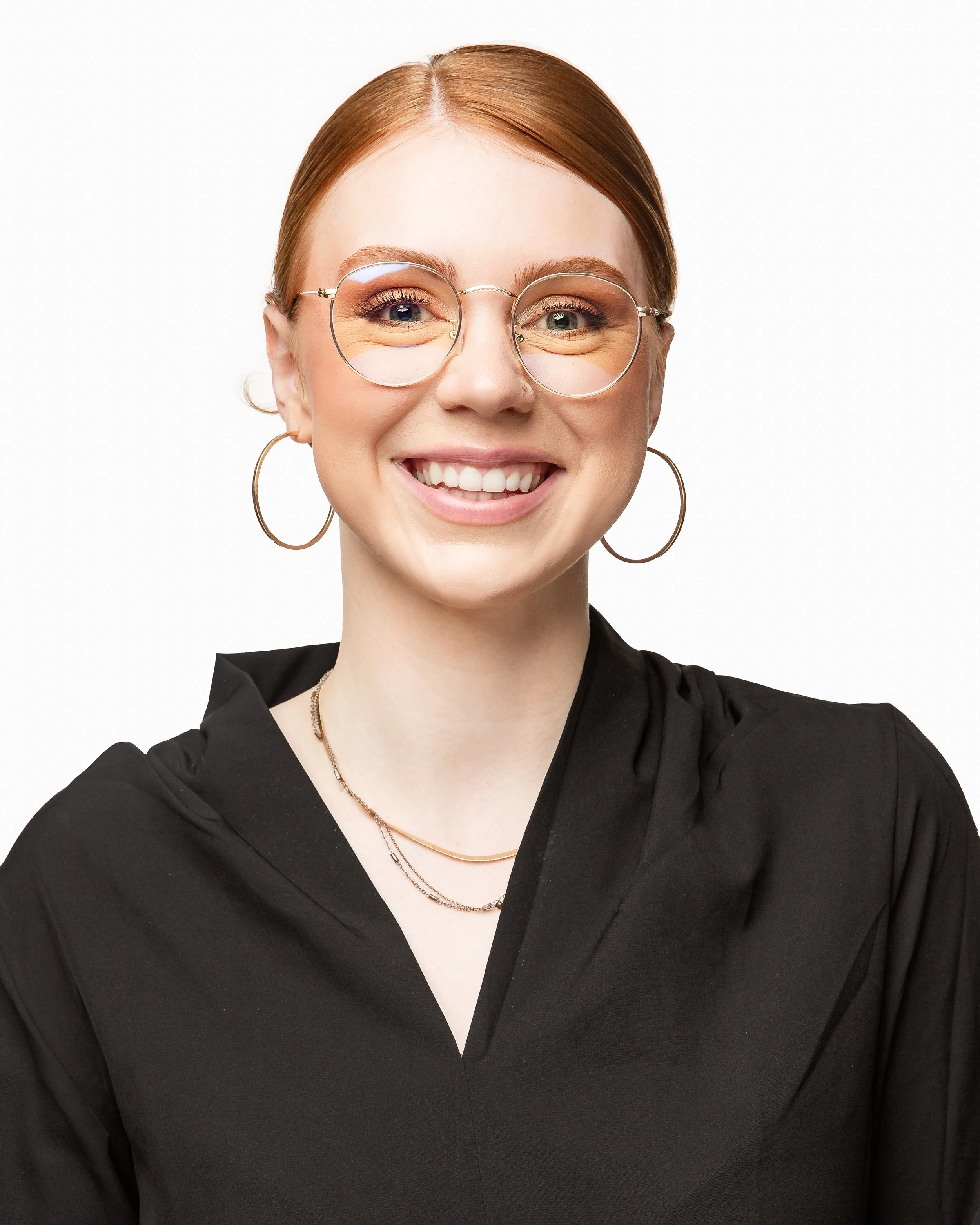 A young woman with fair skin, red hair tied back, wearing round glasses, large hoop earrings, layered necklaces, and a black top, smiling in front of a plain white background.